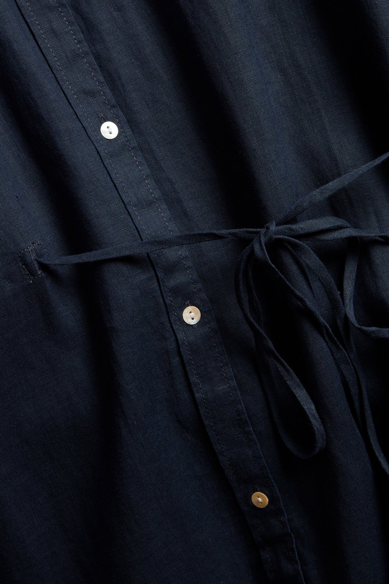 Närbild framifrån på en mörkblå linneskjortklänning med knappslå och knutna midjeband.