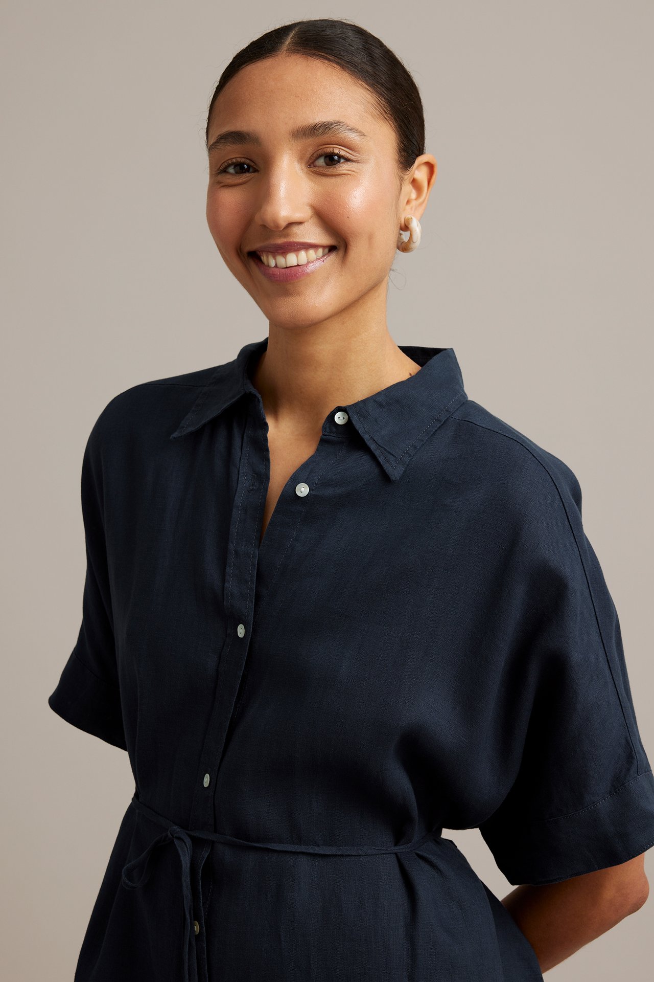Framifrån: Leende kvinna i mörkblå linneskjortklänning med knappar, knyt i midjan. Bär ljusa ringörhängen.