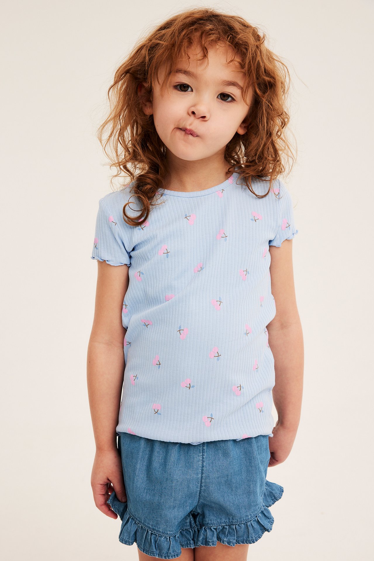 Framifrån: ett barn i ljusa denimshorts med volangkant, tillsammans med en ljusblå mönstrad ribbad t-shirt.