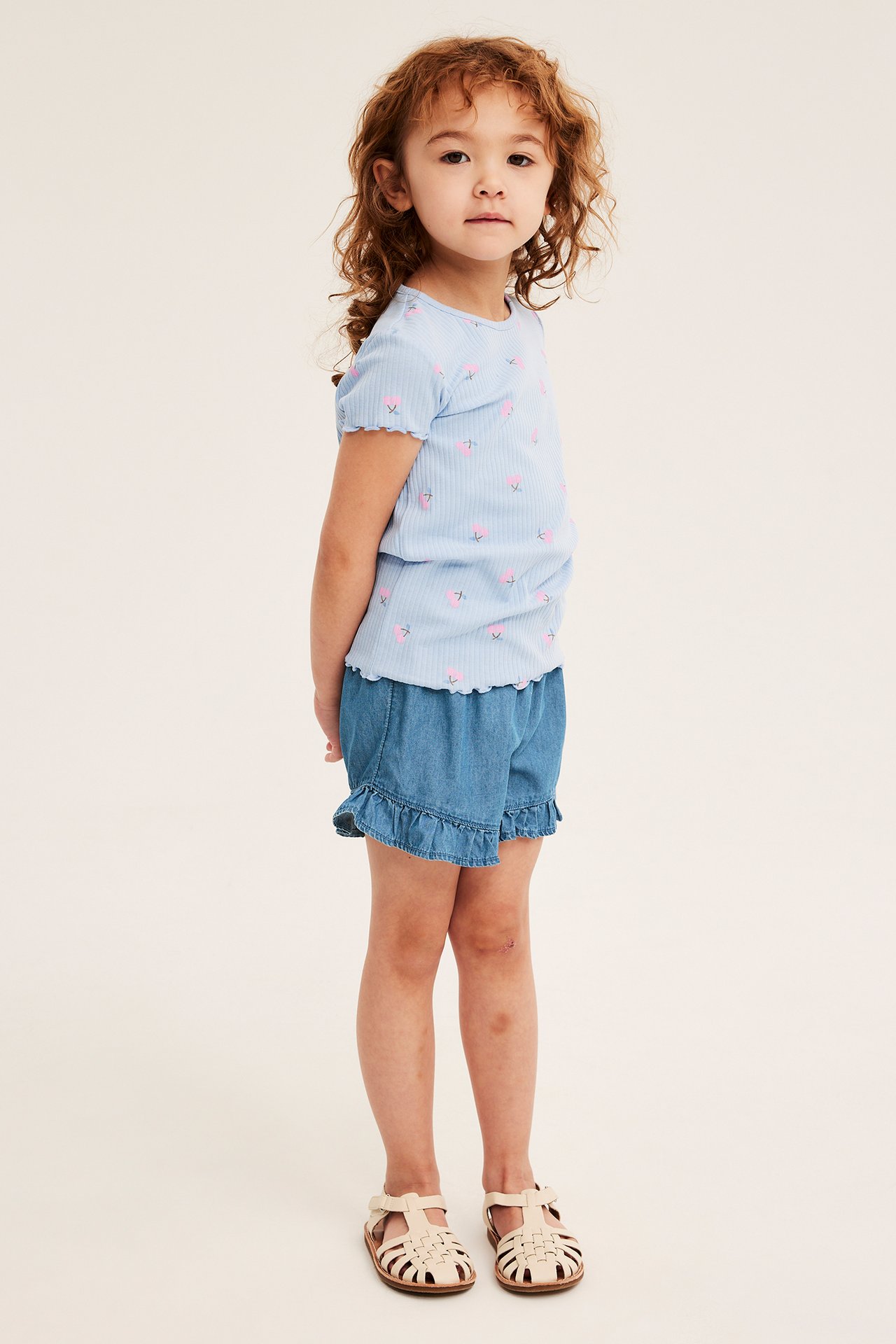 Sidovy av ett barn i ljusa denimshorts med volanger, en ljusblå blommig ribbad t-shirt och beiga sandaler.