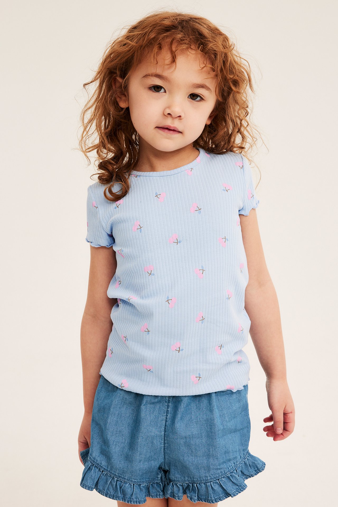 Frontvy: Ett barn bär ljusblå jeansshorts med volangkant och en ljusblå ribbad blommig t-shirt.