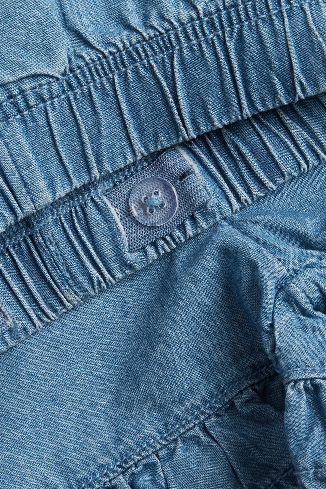 Zbliżenie jasnoniebieskich szortów jeansowych z falbanką, elastyczną talią i regulowanym guzikiem.