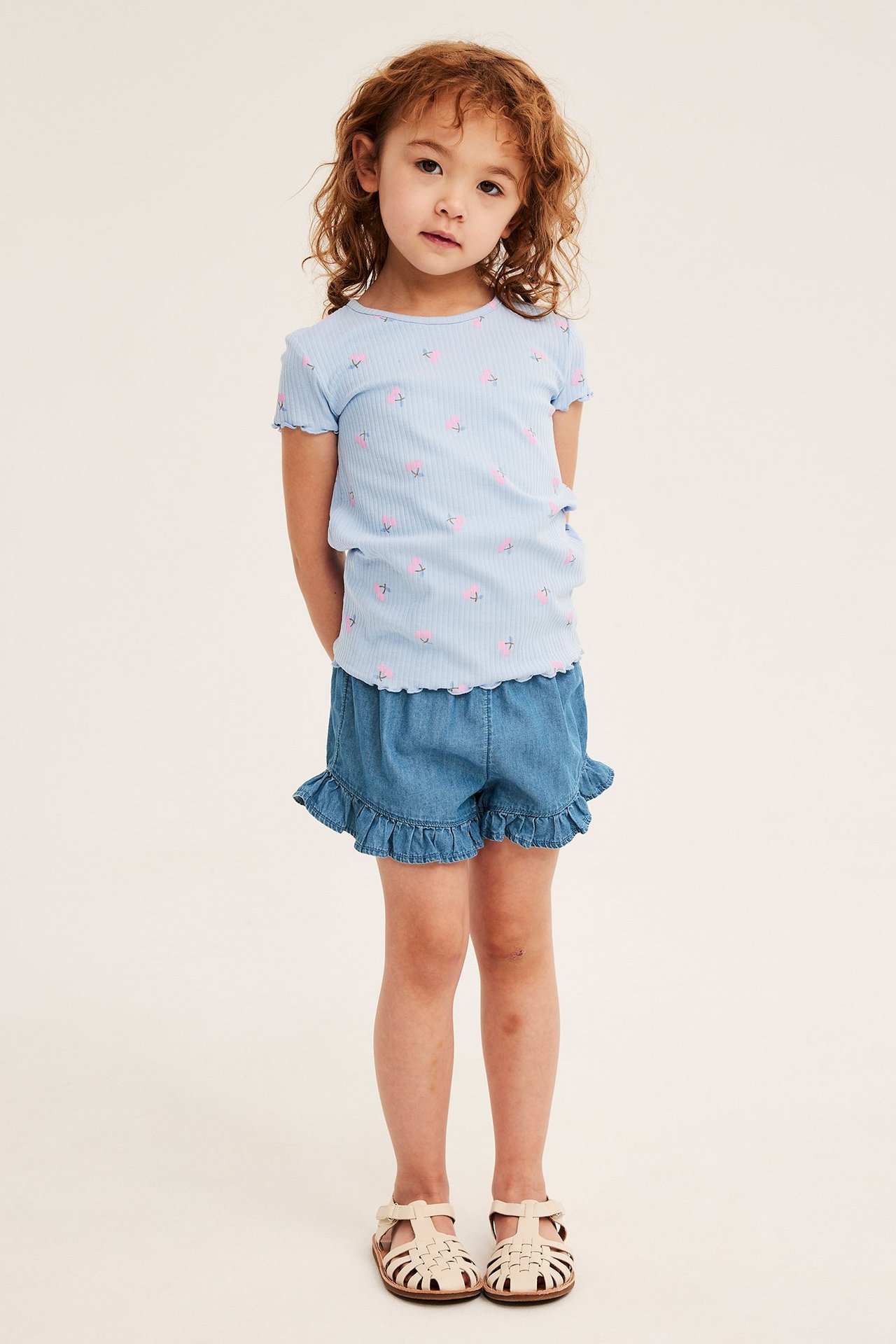 Widok z przodu: Dziecko w jasnoniebieskich jeansowych szortach z falbankami, błękitnej koszulce w truskawki i beżowych sandałach.