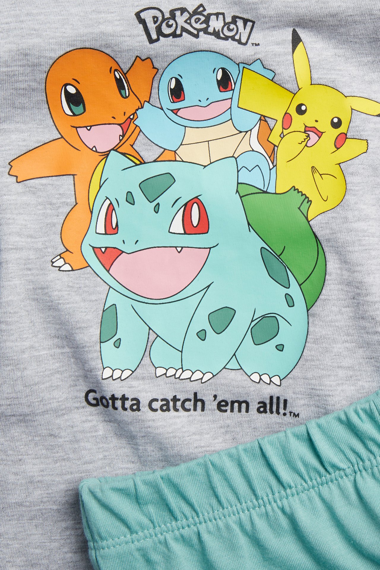 Närbild framifrån: Barns ljusgrå pyjamasset i bomull med Pokémon-tryckt topp och turkosa shorts.