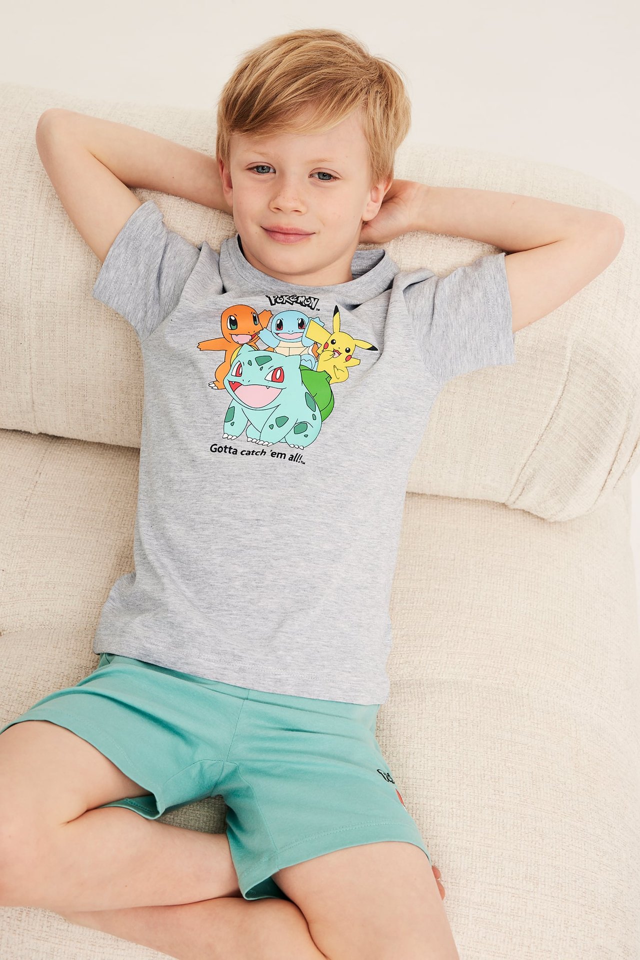 Poika harmaa Pokémon-T-paita ja turkoosit shortsit, makailee sohvalla kädet takana.