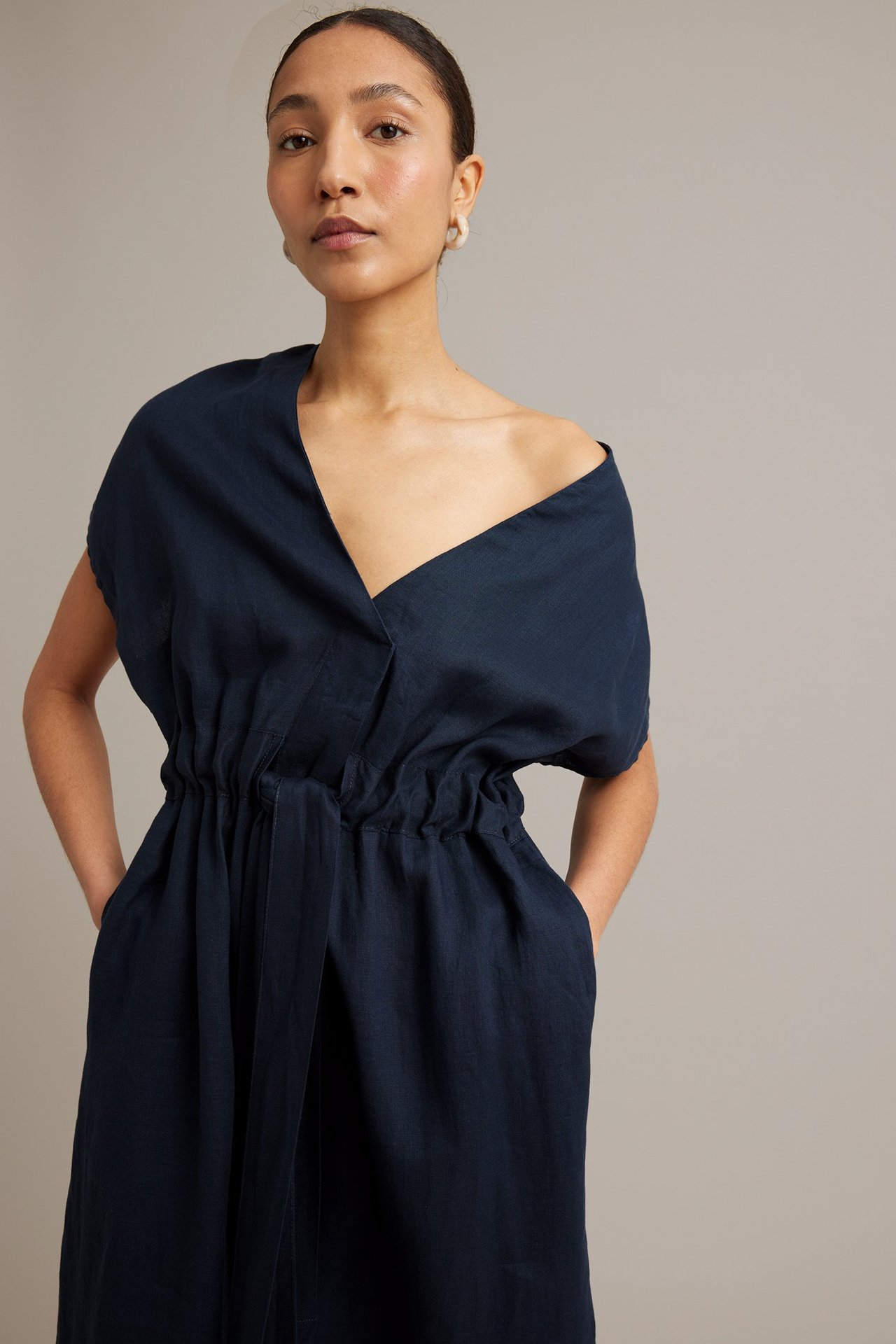 Framifrån: kvinna i mörkblå linneklänning med V-ringning, midjeband, sidfickor och örhängen.