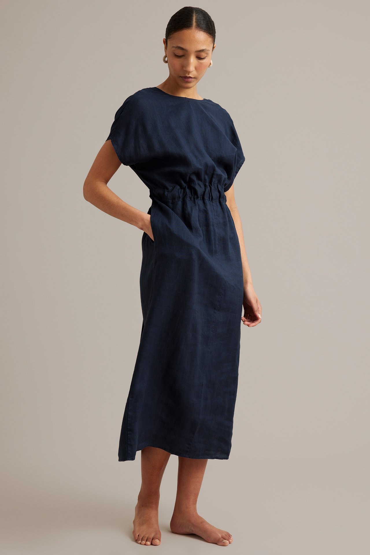 Sidovy av kvinna i mörkblå linneklänning med korta ärmar, resår i midjan och sidfickor.