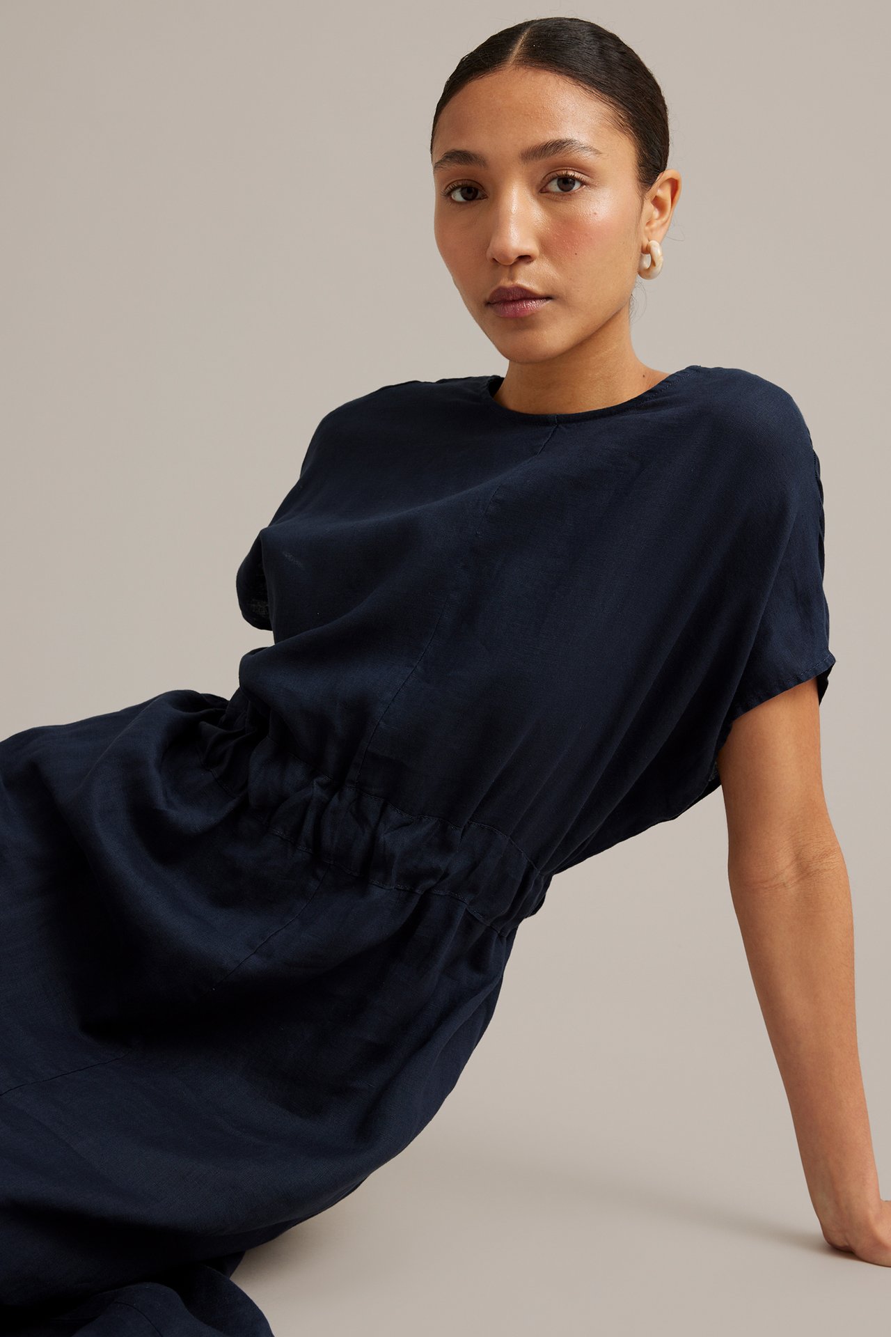 Kvinna i mörkblå linneklänning med rund hals, resår i midjan. Hon bär ljusa ringörhängen.