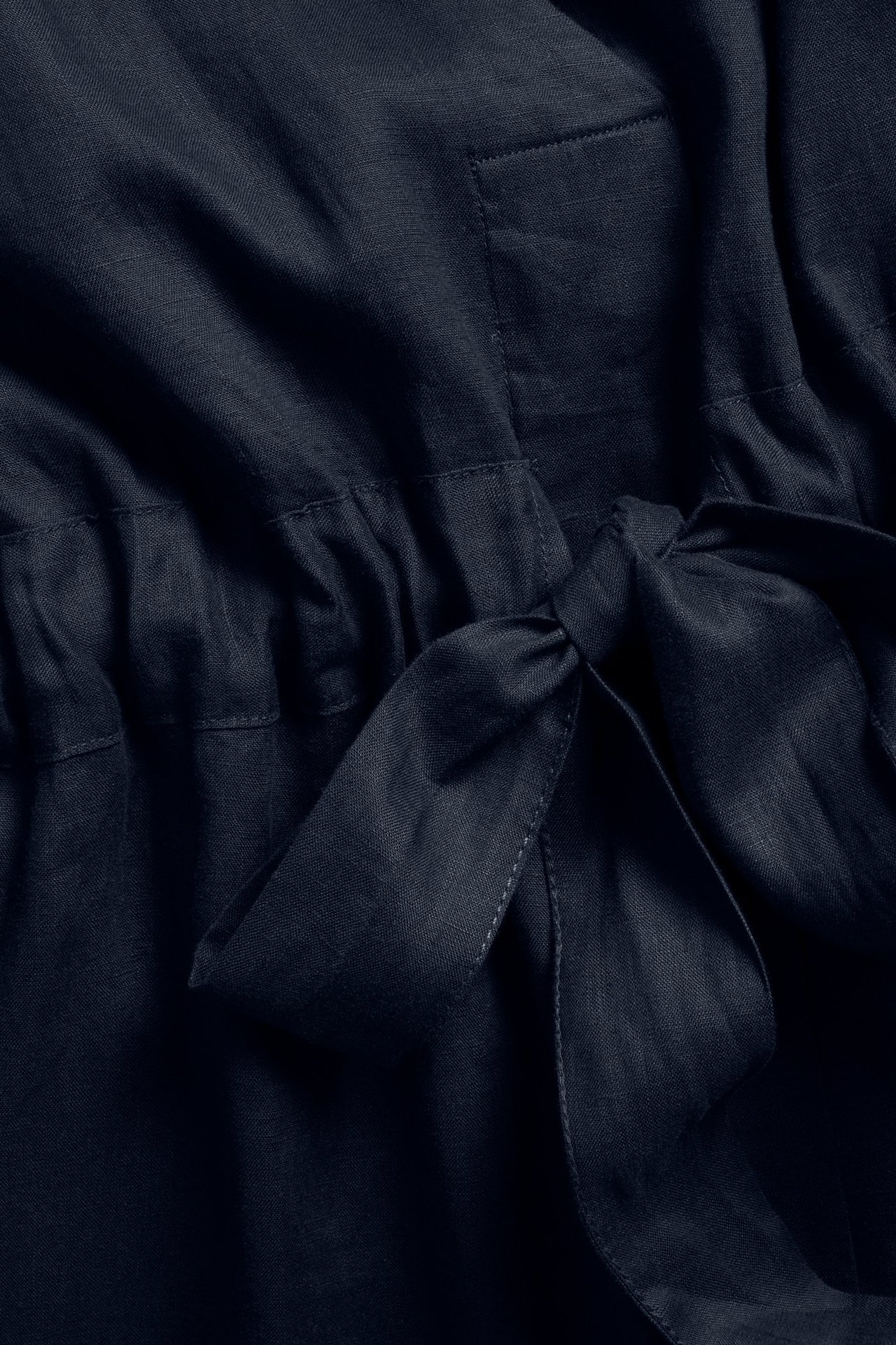 Närbild framifrån på en mörkblå linneklänning med rynkad midja och knutet knytband.