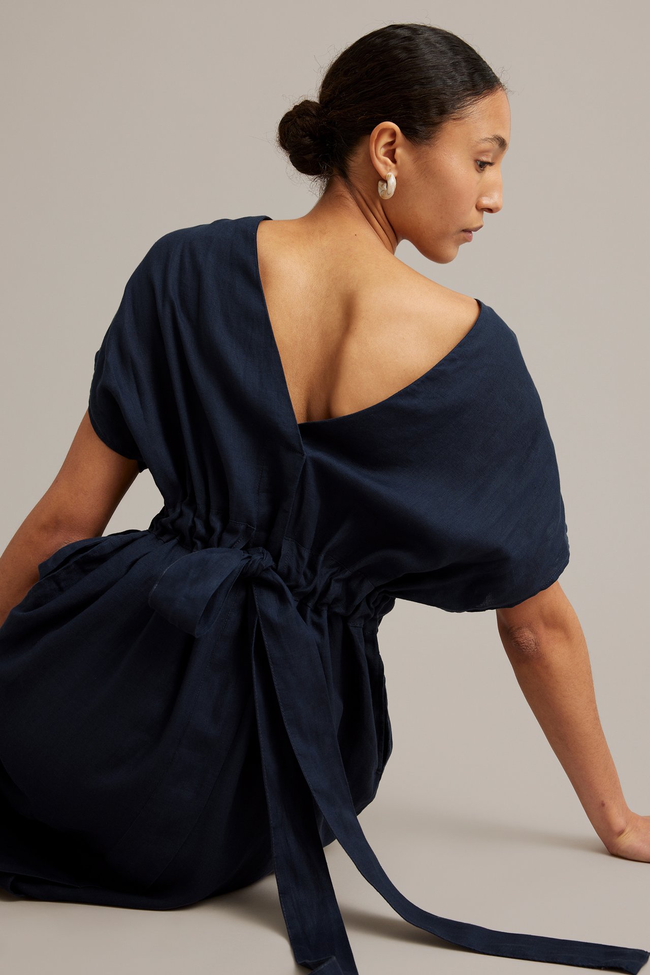 Bakifrån: kvinna i mörkblå linneklänning, djup V-ringning, knuten resår i midjan, vita ringörhängen.