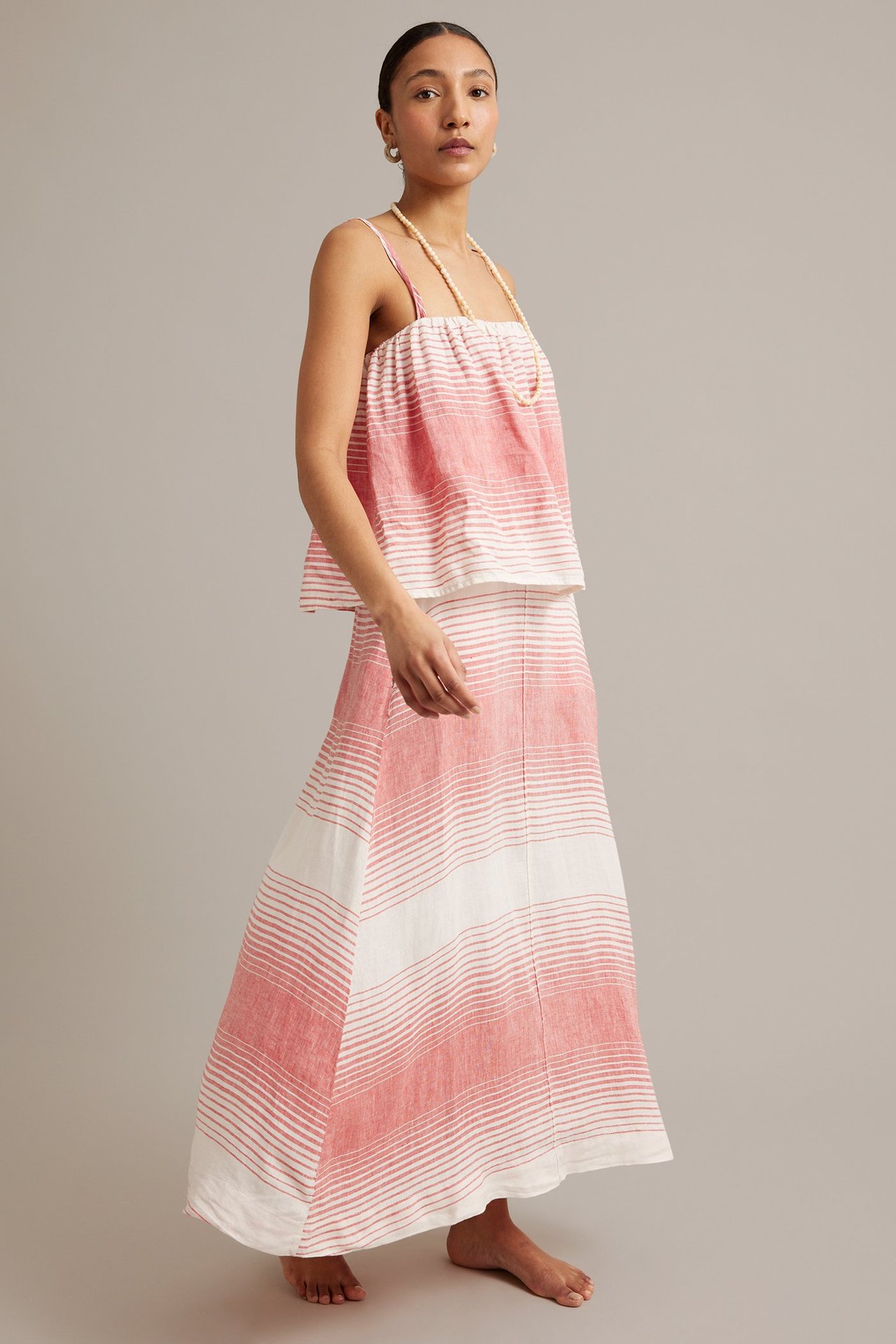 Framifrån en kvinna i röd och vitrandig lång linnekjol och matchande linne, med pärlhalsband.