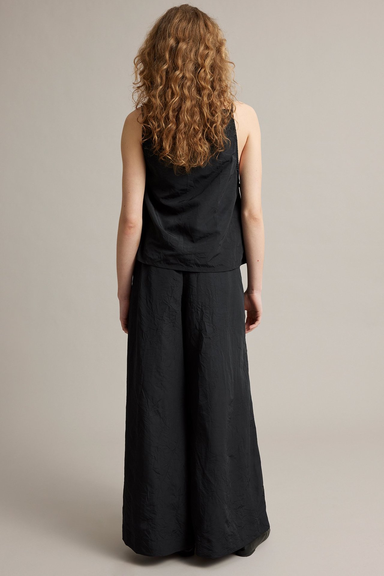 Widok z tyłu kobiety w czarnych, szerokich spodniach ze strukturą materiału, w połączeniu z czarnym topem bez rękawów.