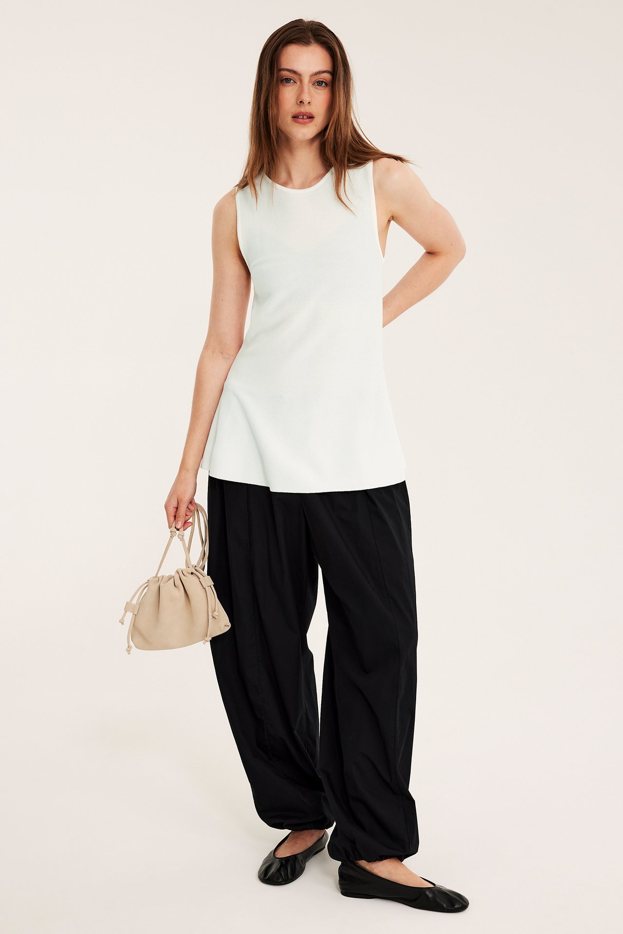 Front view: Kvinna i offwhite stickad peplumtopp, svarta vida byxor, svarta ballerinaskor, håller beige handväska.