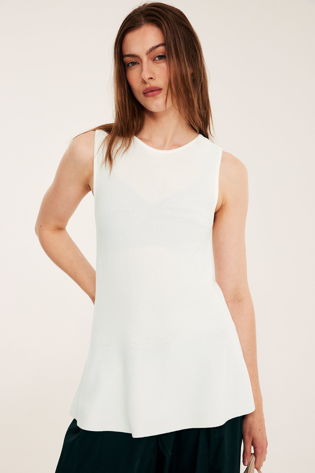 Framifrån: Kvinna i offwhite stickad peplumtopp och mörkgröna byxor, håller en ljus väska.