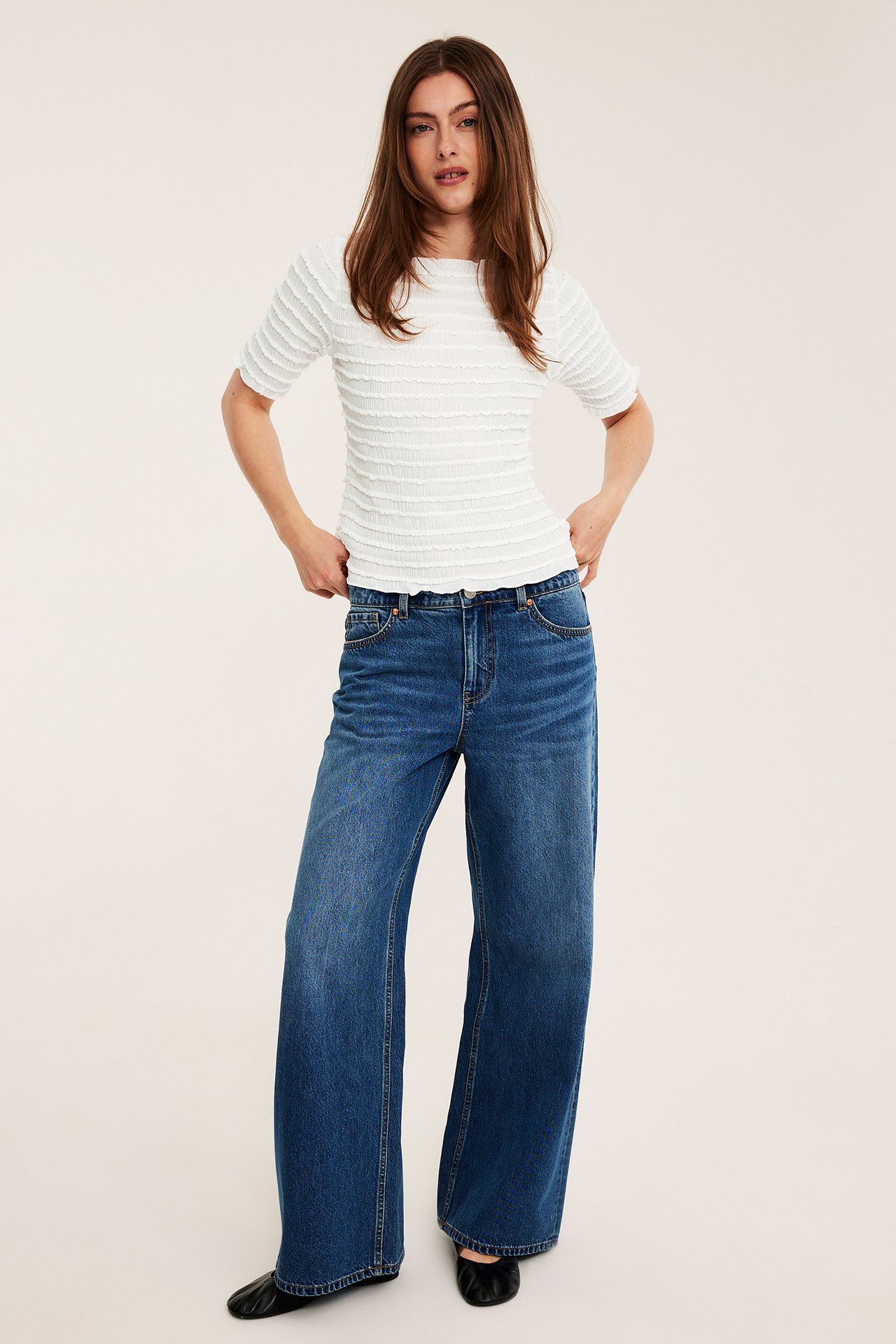 Framifrån en kvinna i offwhite strukturerad kortärmad båtringad topp, blåa vida jeans och svarta platta skor.