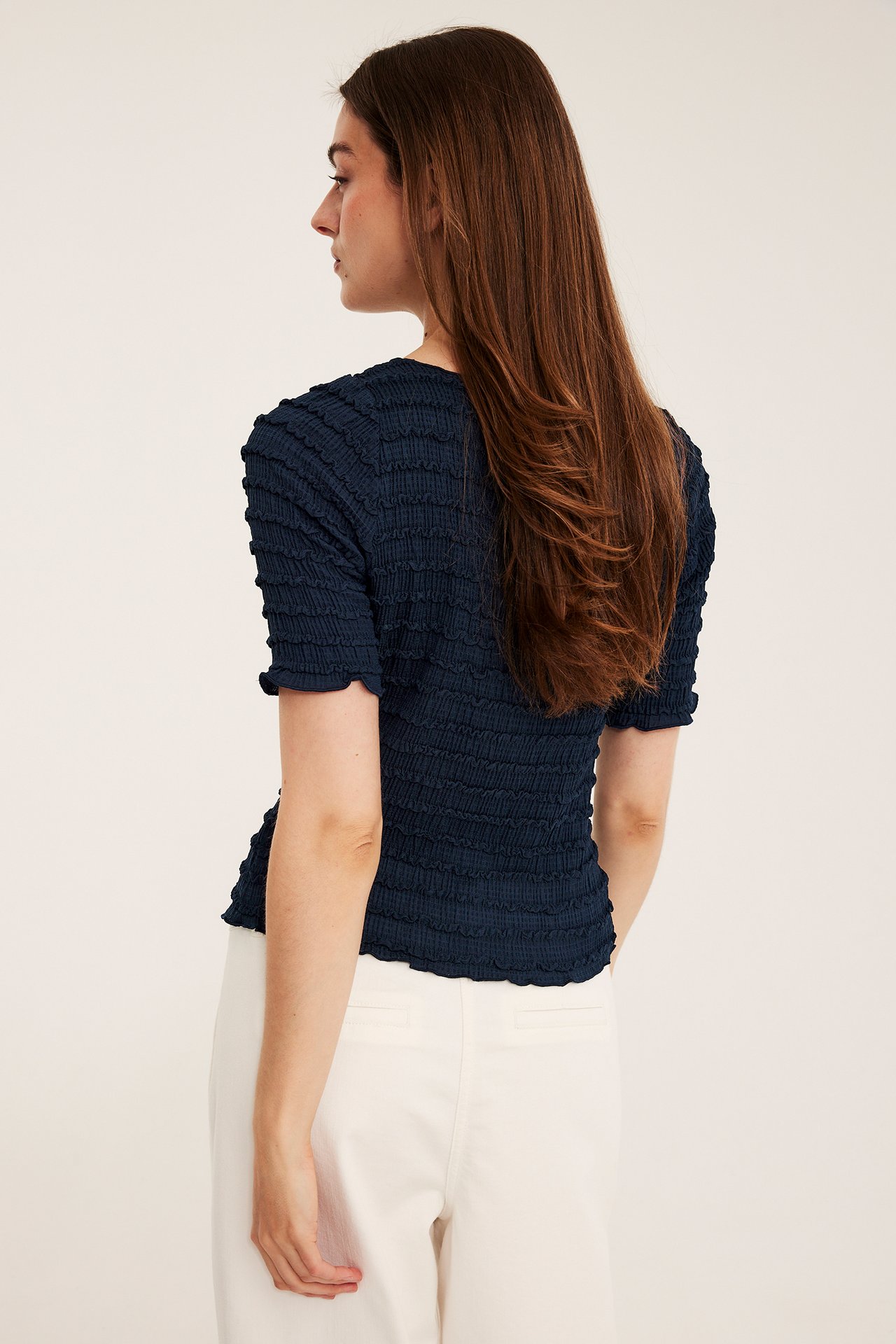 Widok z boku i tyłu kobiety w granatowym, teksturowanym topie z dekoltem łódkowym i krótkimi rękawami, oraz białych spodniach.