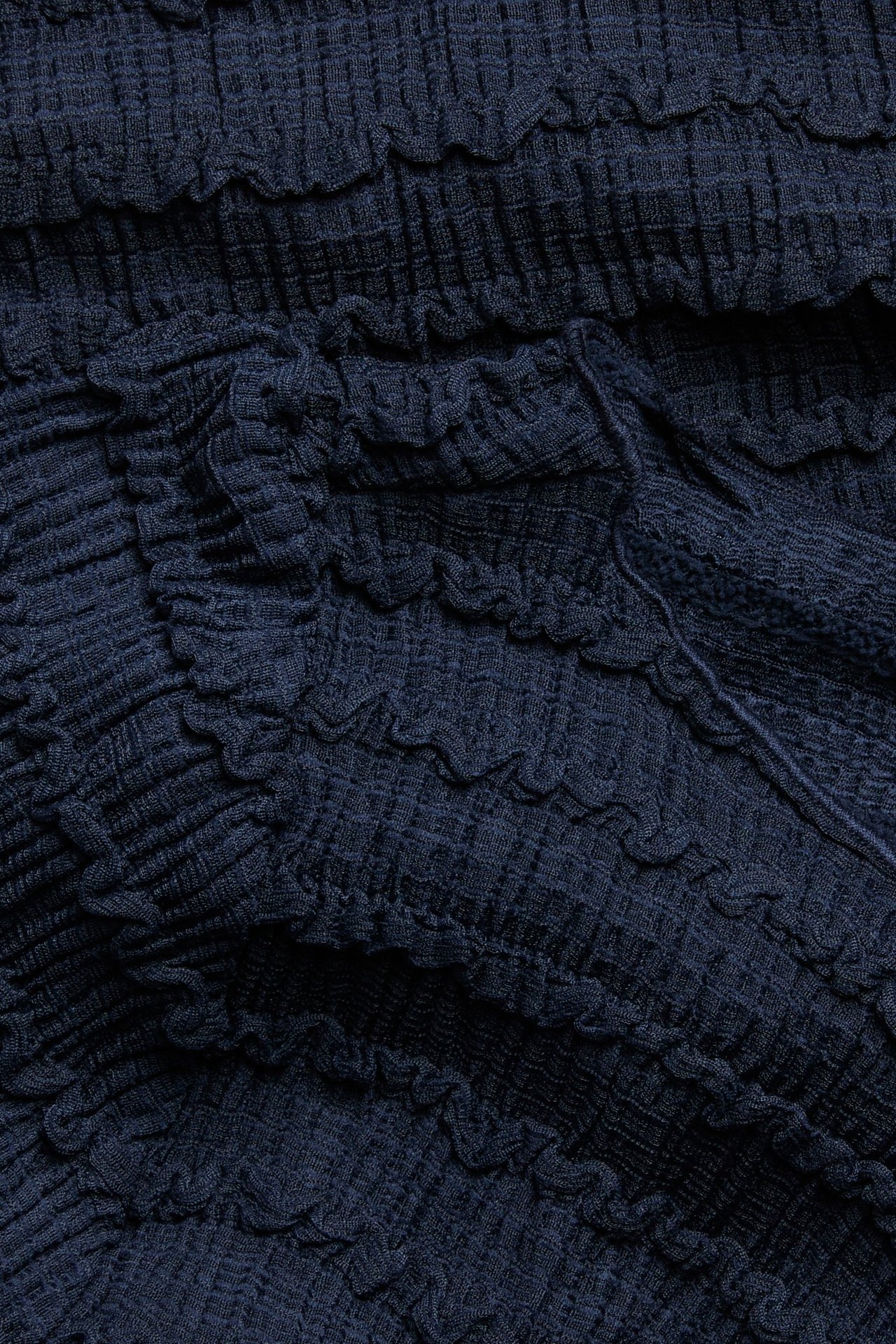 Närbild av mörkblått strukturerat topptyg med krusiga och volangdetaljer.