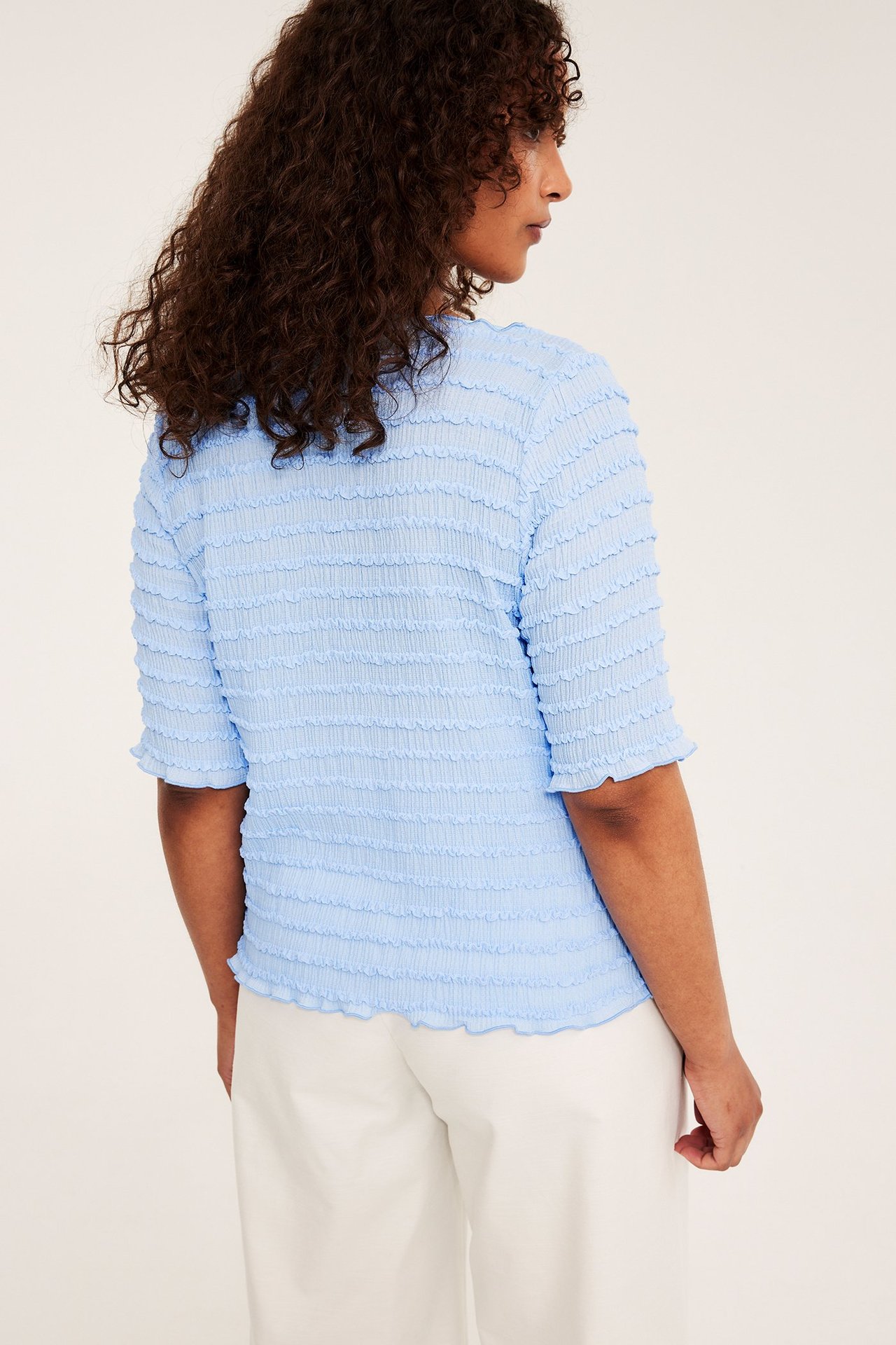 Takasivu naisesta, jolla on vaaleansininen strukturoitu venepääntie-toppi ja valkoiset leveät housut.