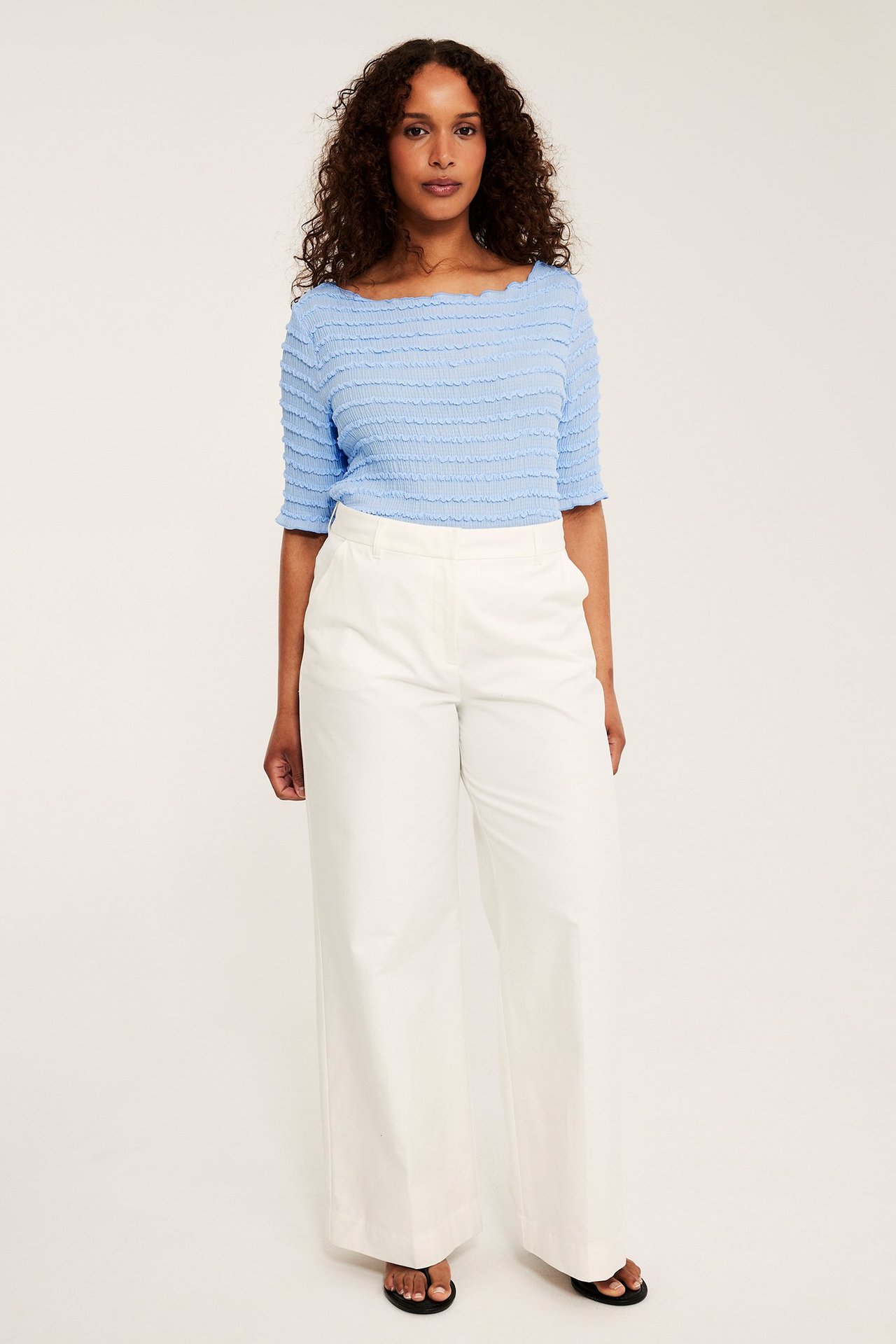 Framsida: Kvinna i ljusblå strukturerad båthalsad topp, vita vida byxor och svarta flip-flops.