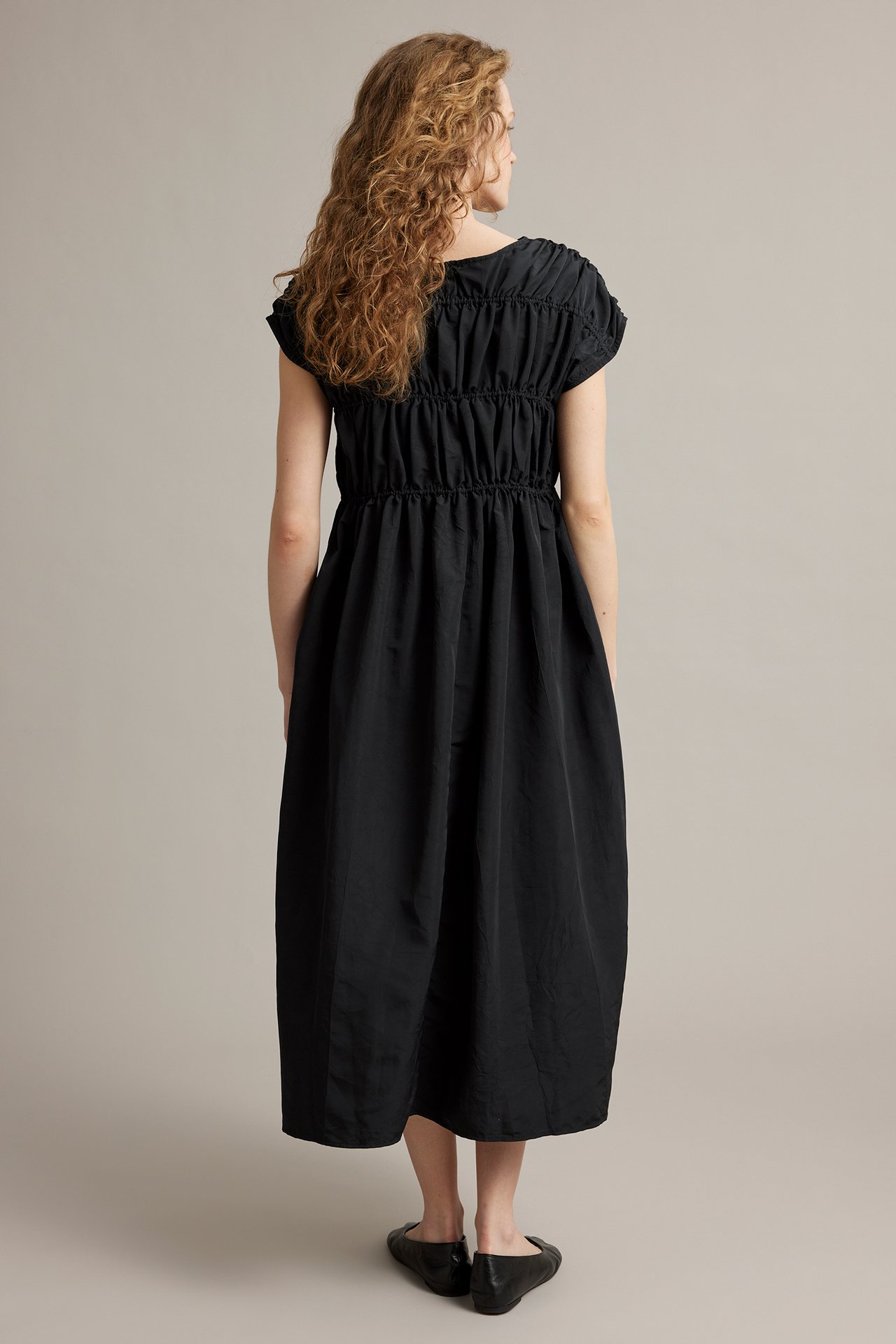 Naisen takaa näkymä, yllään musta polyesteri midimekko rypytyksillä ja mustat ballerinat.