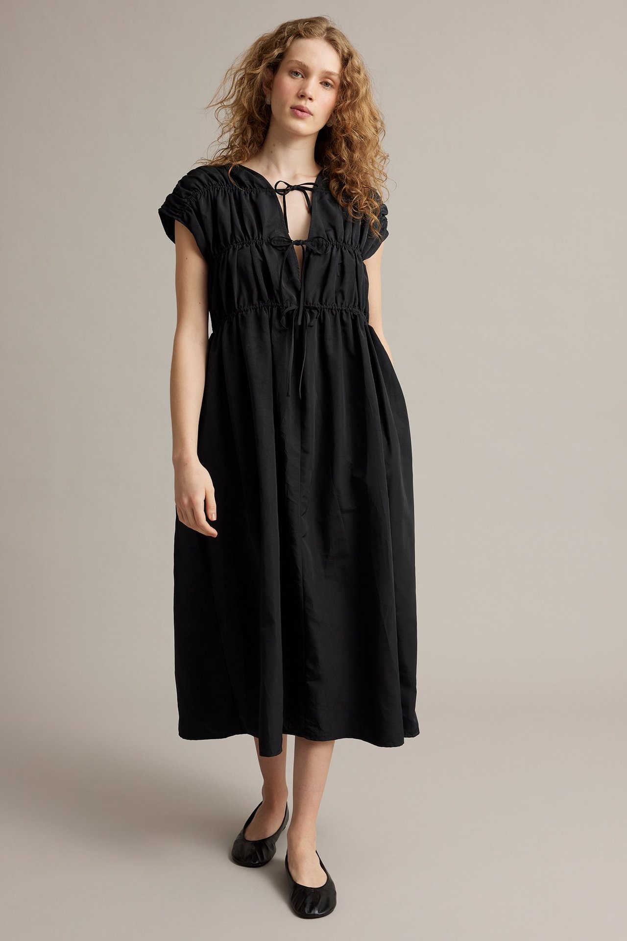Forfra: kvinne i svart polyester-midikjole med rynket knyteoverdel og svarte ballerinasko.