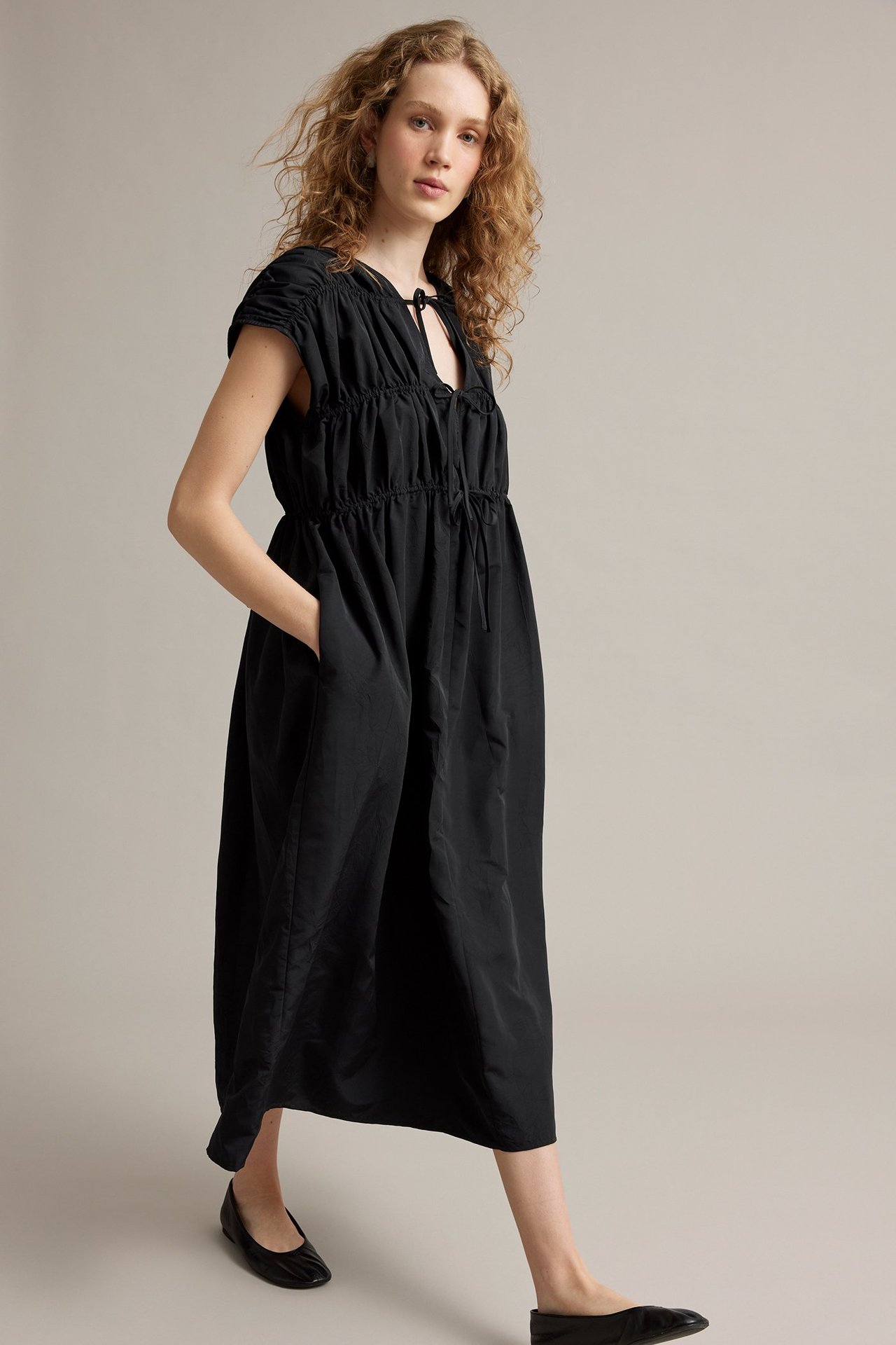Etunäkymä, nainen mustassa pitkässä polyesterimekossa, jossa solmittavat yksityiskohdat ja rypytetty yläosa, jalassa mustat ballerinat.