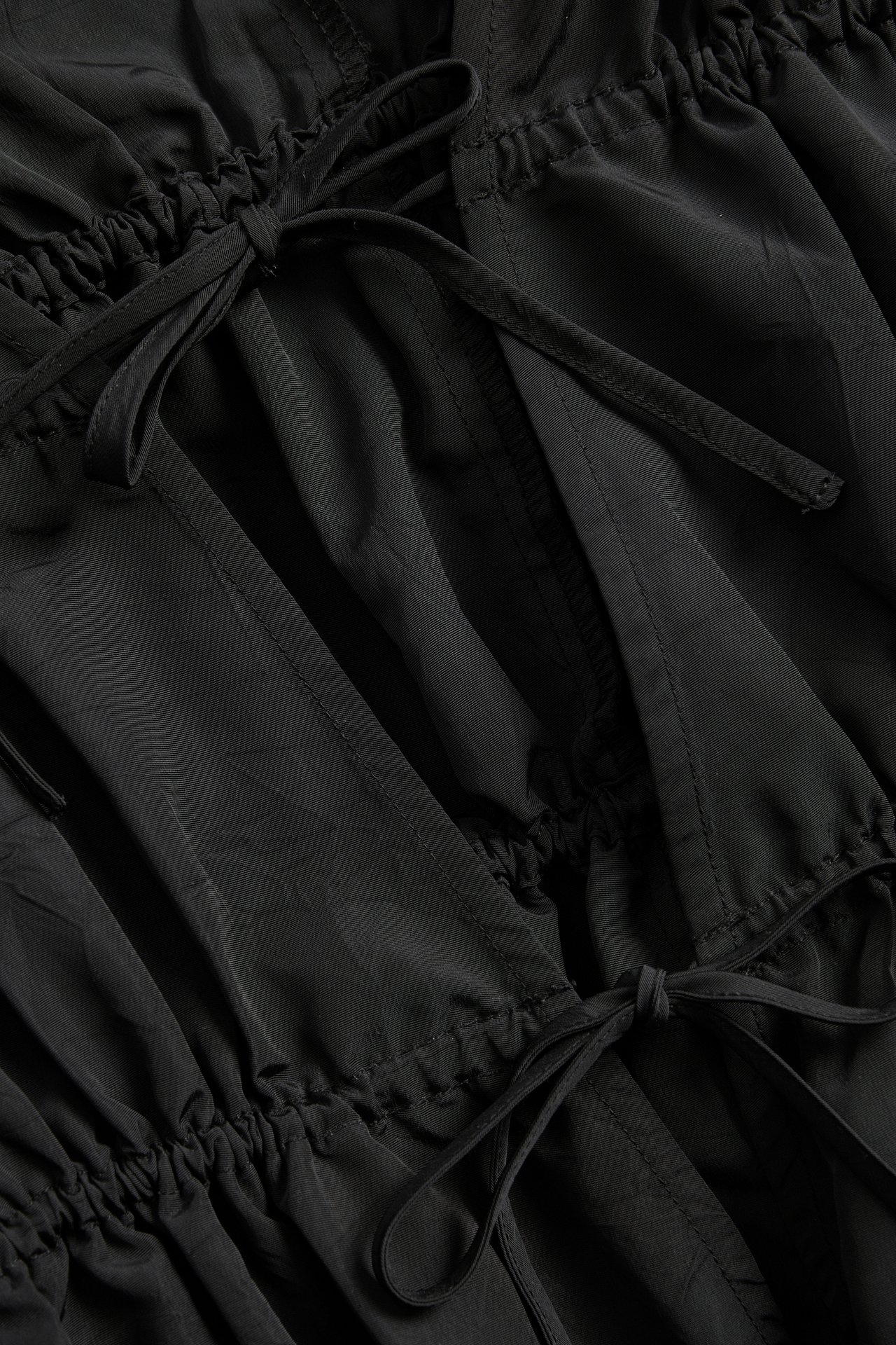 Närbild på en svart polyesterklänning med strukturerad yta, rynkade kanaler och två knytband.