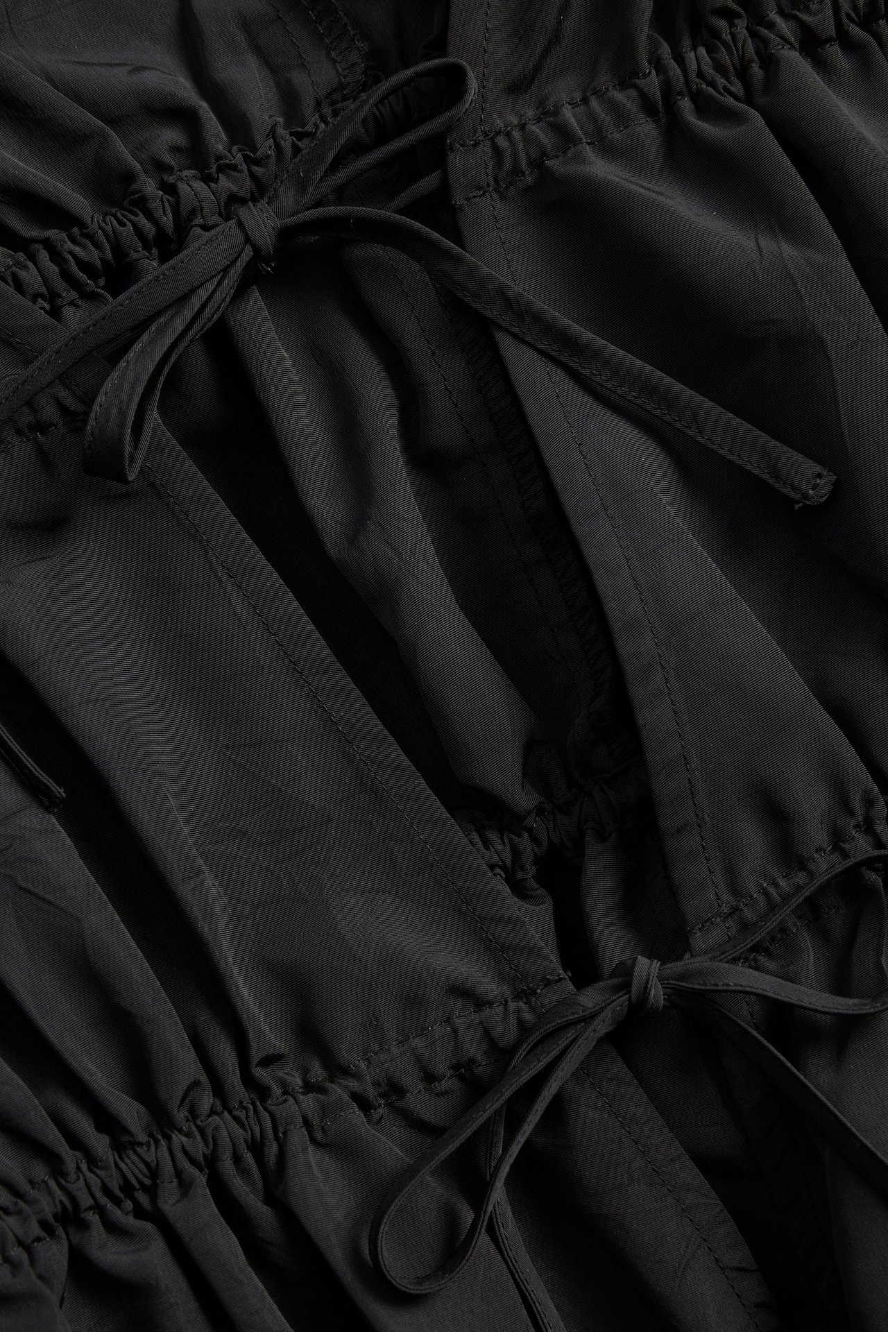 Nærbilde av en svart polyesterkjole med strukturert overflate, rynkede kanaler og to knytebånd.