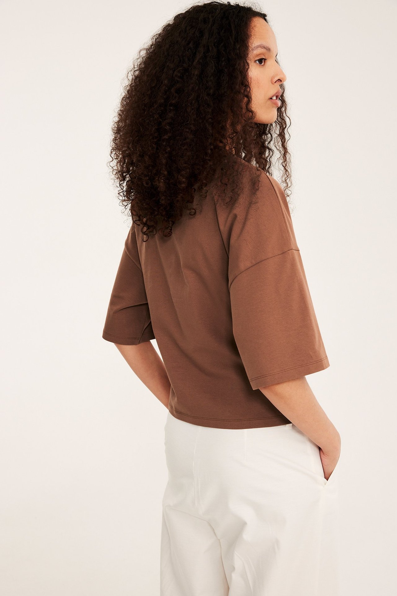 Widok z boku kobiety: brązowy dopasowany t-shirt z obniżoną linią ramion i białe spodnie.