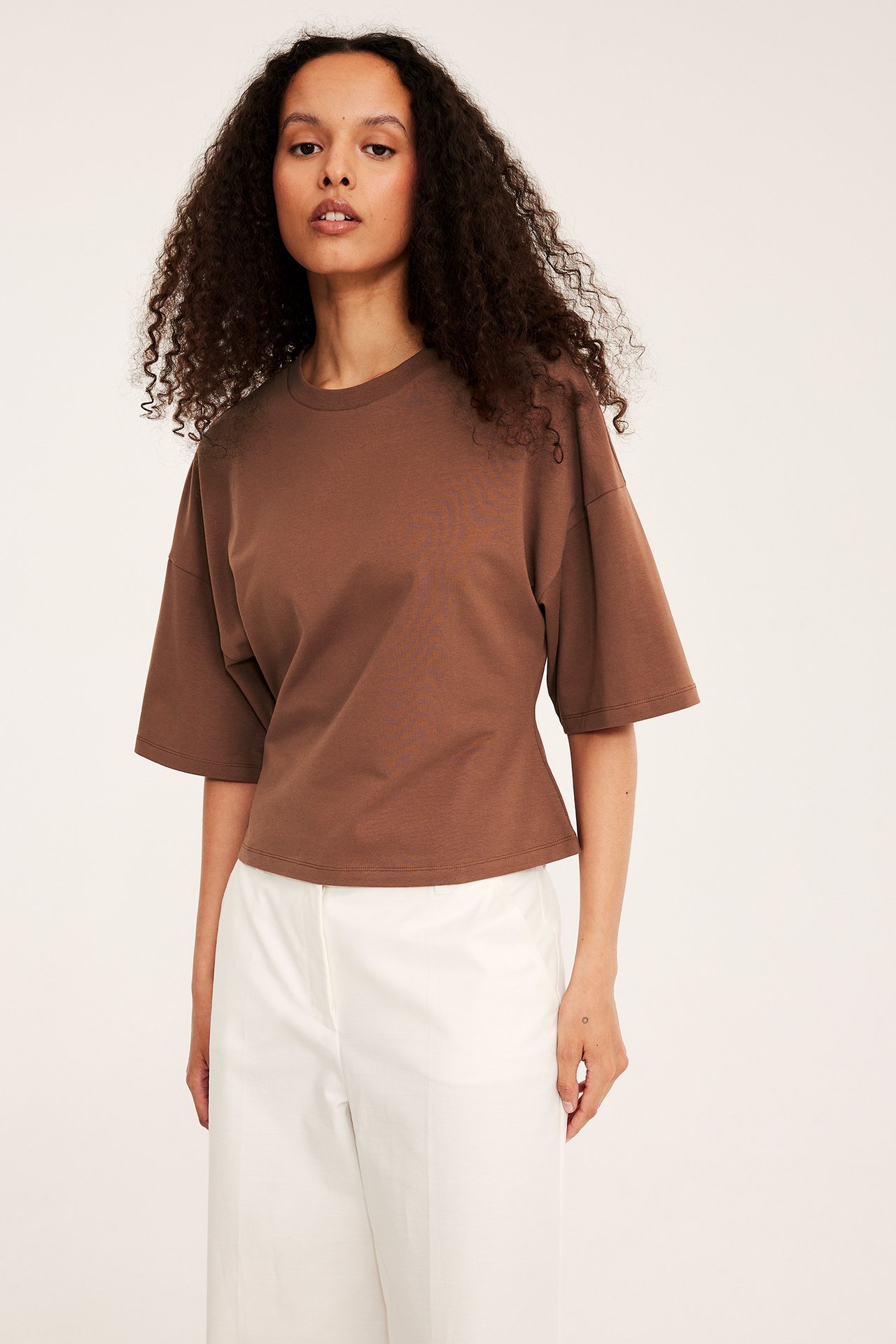 Widok z przodu: kobieta w brązowym dopasowanym t-shircie z opuszczonymi ramionami i białych spodniach.