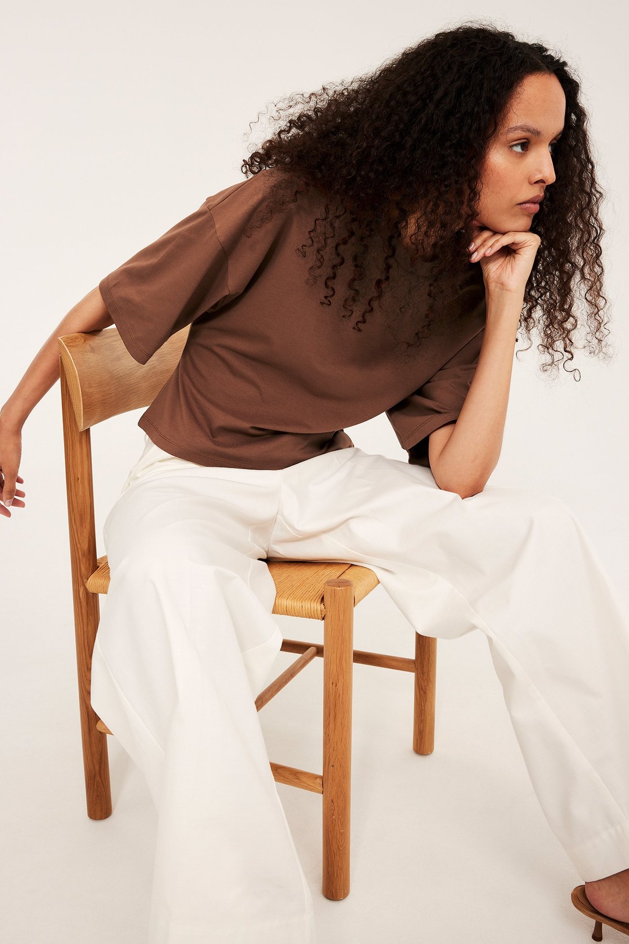 Widok z przodu: kobieta w brązowej, dopasowanej koszulce z opuszczonymi ramionami i białych spodniach.