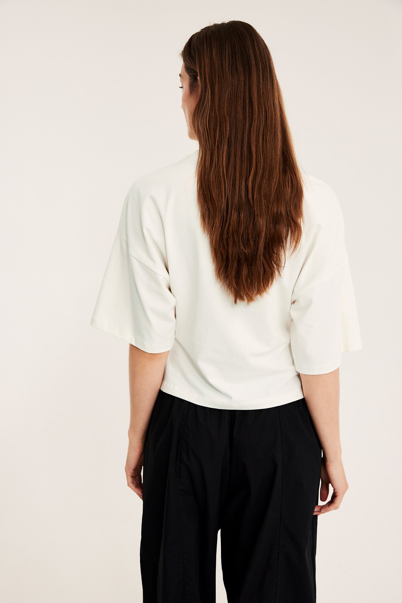 Widok z tyłu kobiety w dopasowanym T-shircie ecru z obniżoną linią ramion i luźnych czarnych spodniach.