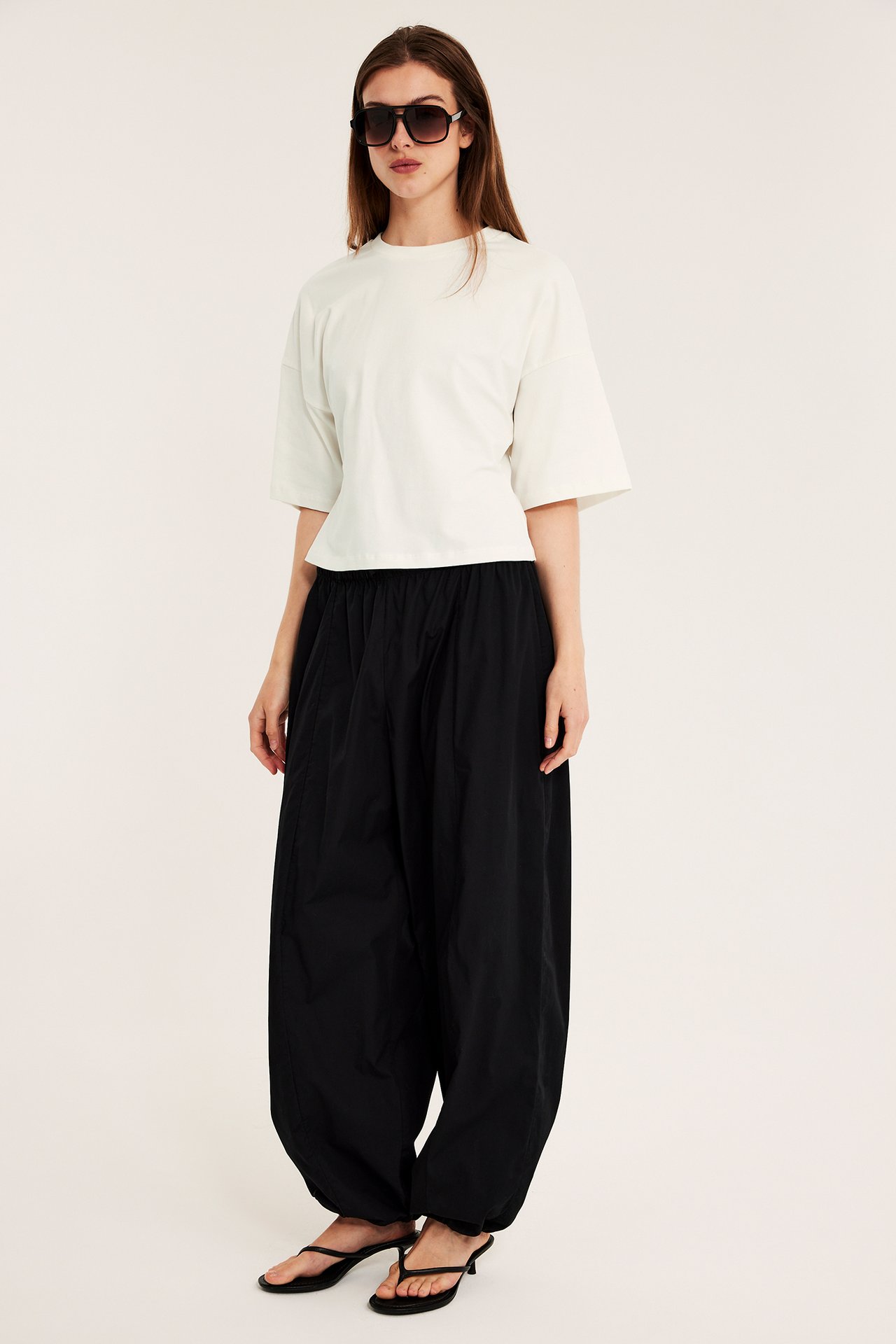 Kobieta w off-white dopasowanym T-shircie, czarnych szerokich spodniach, czarnych sandałach na obcasie i okularach przeciwsłonecznych.