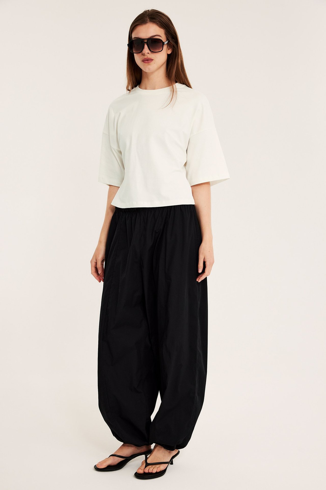 Framifrån, kvinna i figurnära offwhite t-shirt, svarta vida byxor, svarta sandaler med klack och solglasögon.