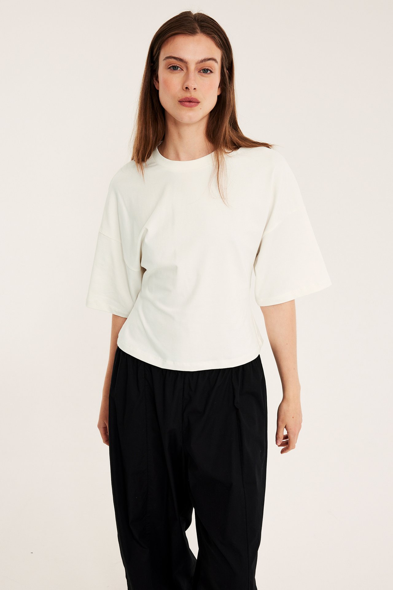 Z przodu: kobieta w dopasowanym, ecru t-shircie z opuszczonymi ramionami i czarnych luźnych spodniach.