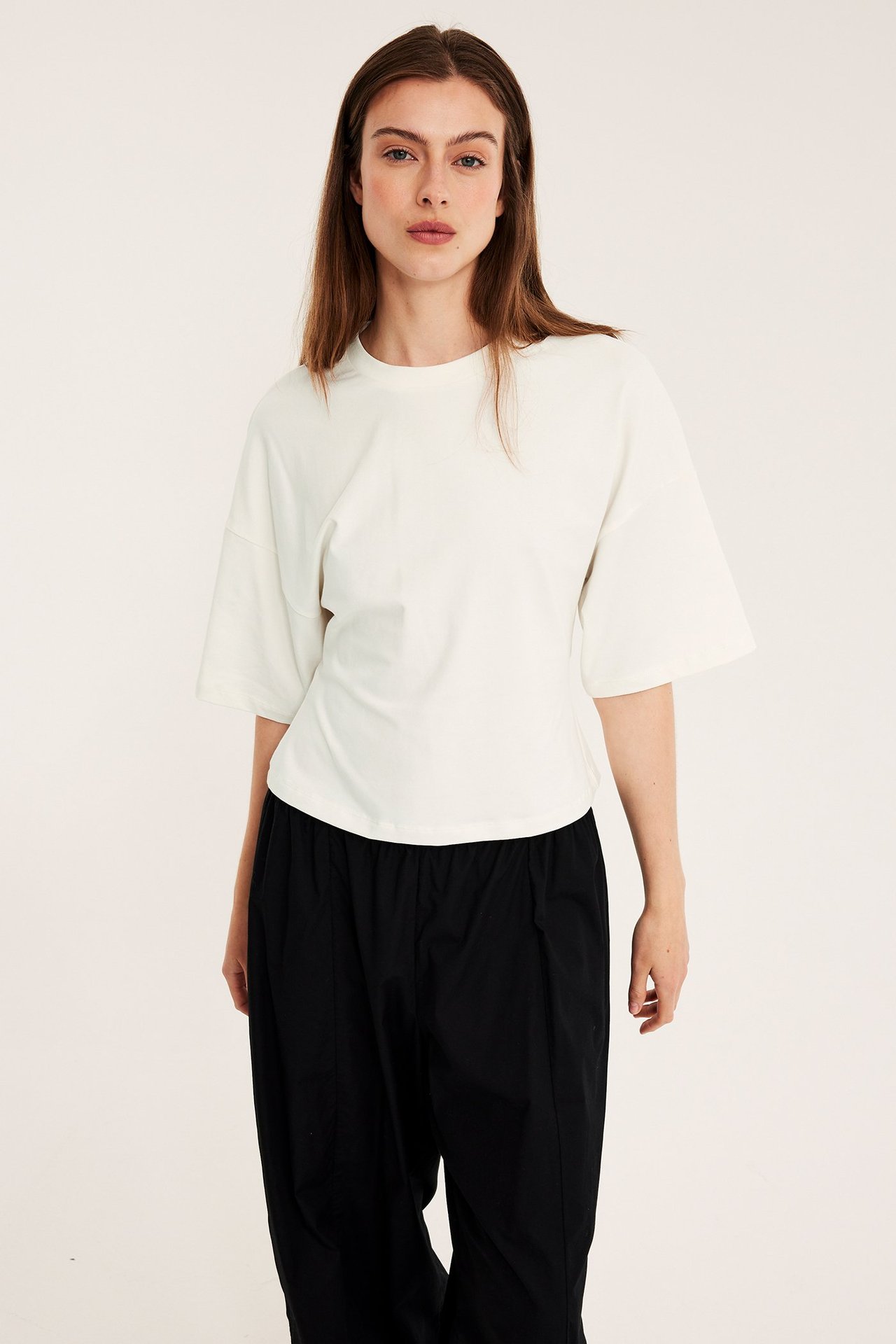 Frontvy av en kvinna i en figurnära offwhite t-shirt med droppade axlar och svarta lösa byxor.
