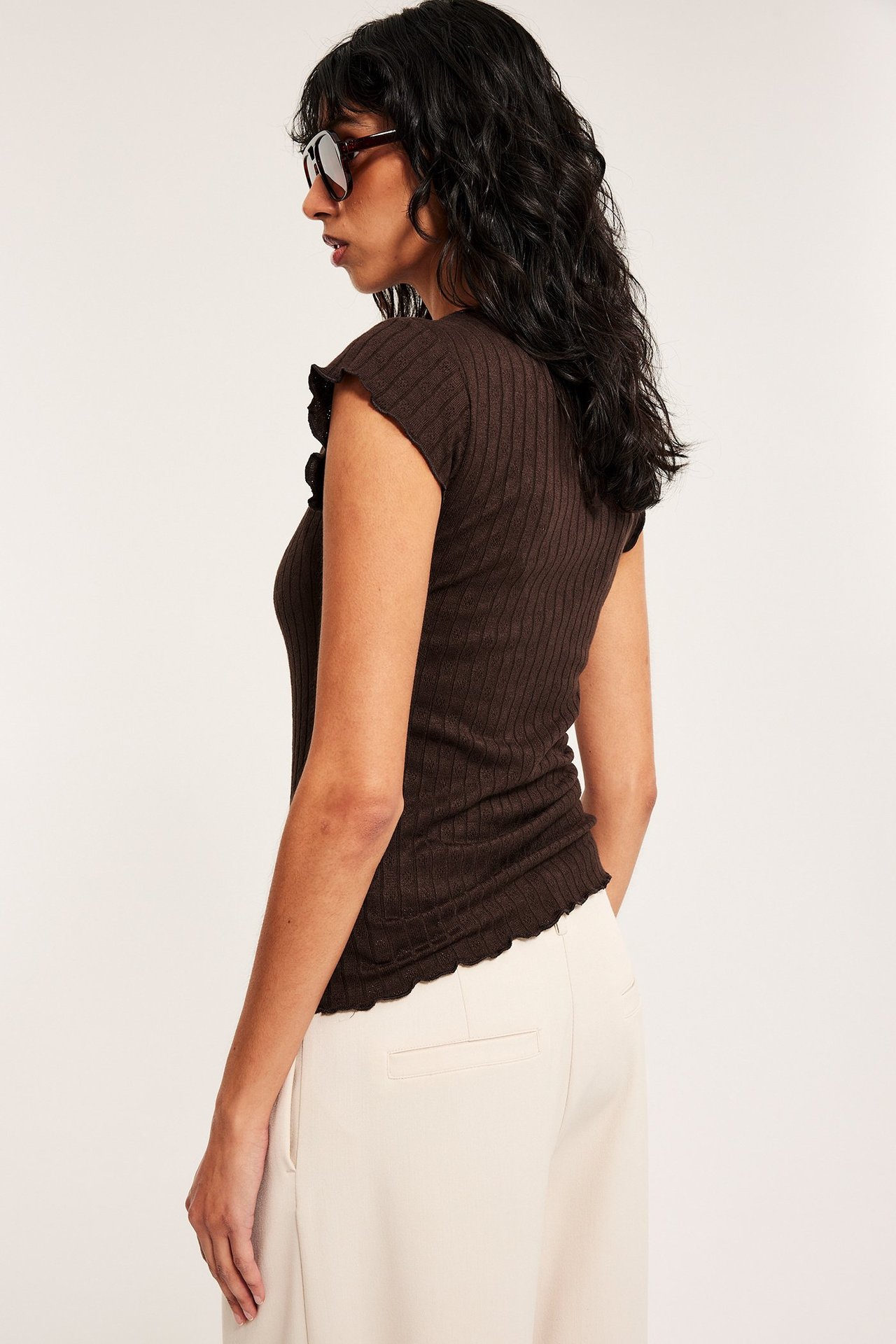 Sideprofil av kvinne i mørk brun ribbet pointelle babytopp, lys beige bukse og solbriller.