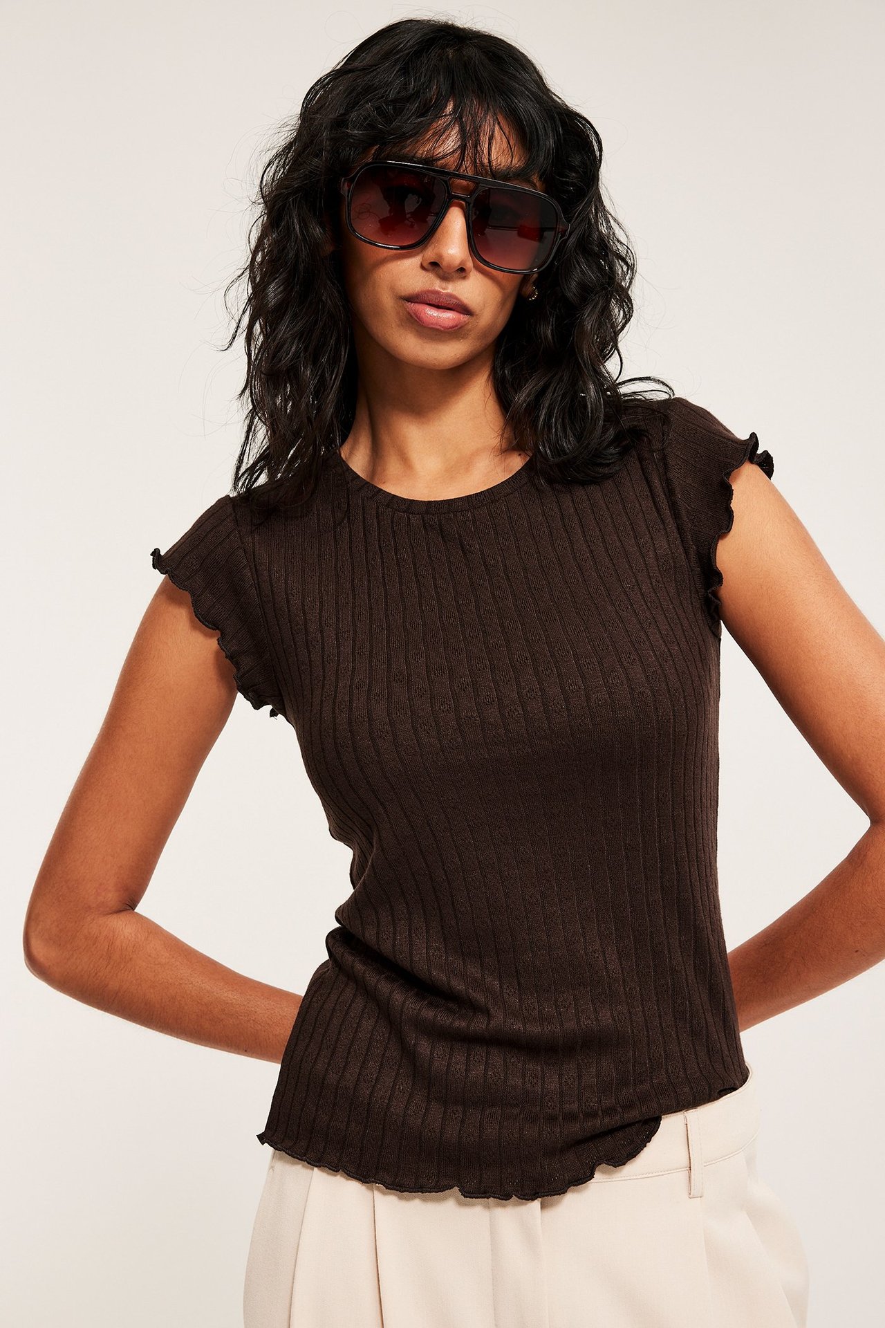 Forfra: Kvinne i mørkebrun ribbet pointelle baby-topp, lyse beige bukser og mørke solbriller.