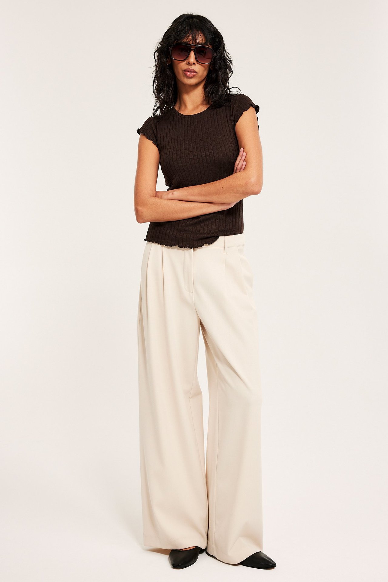 Forfra: Kvinne i mørkebrun pointelle baby tee, beige vide bukser og mørke solbriller.