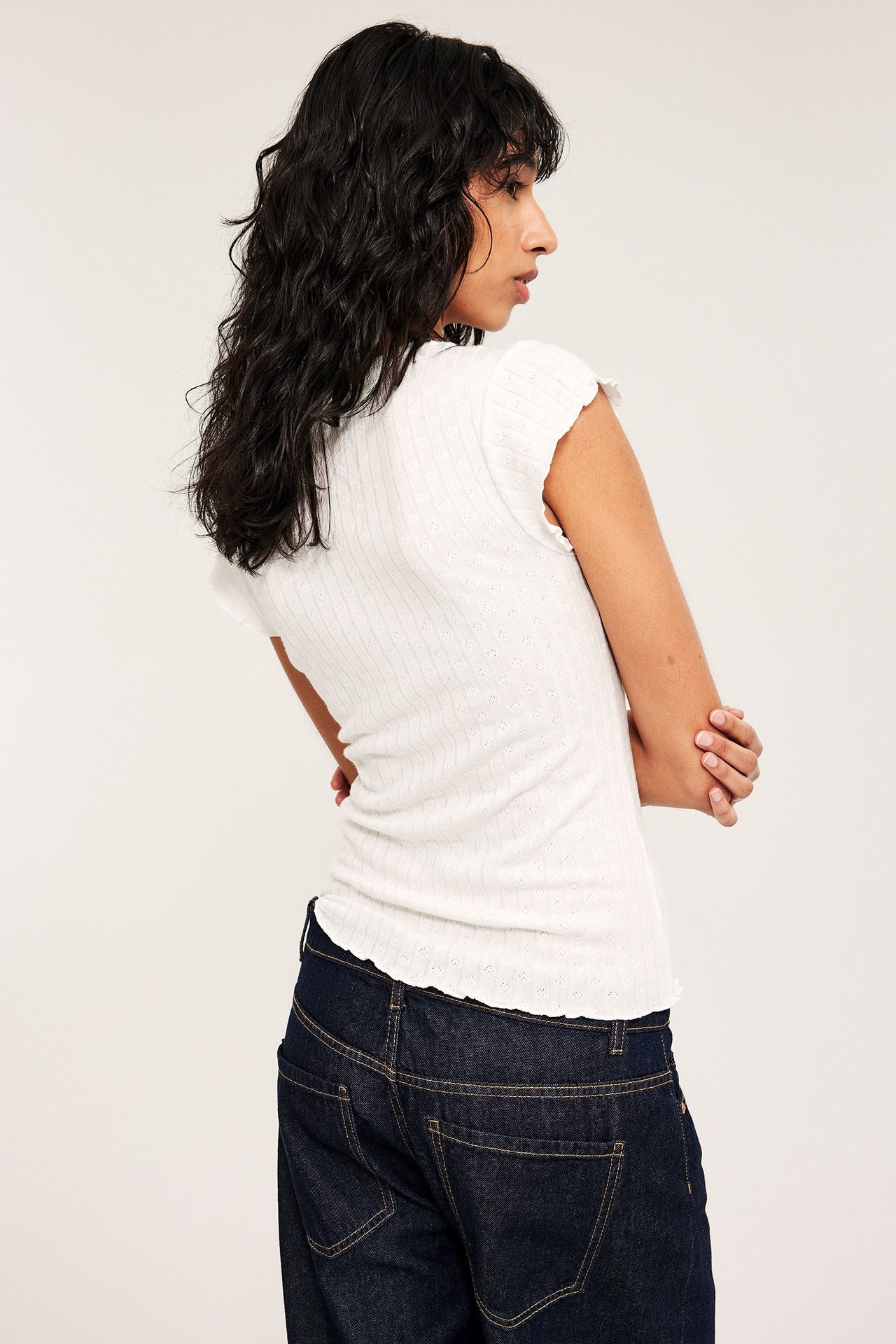 Sidebilde av en kvinne i en offwhite pointelle babytopp med volangkanter og mørkeblå jeans.