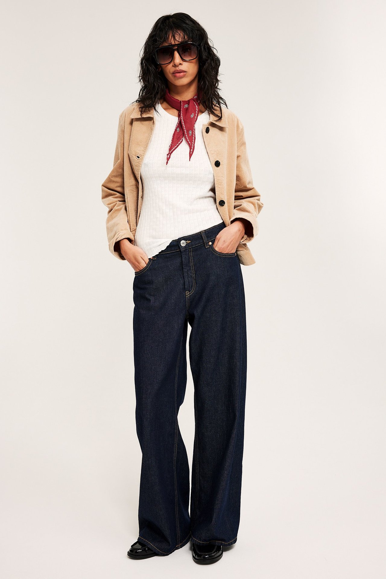 Kvinne i offwhite pointelle baby-topp, beige cordfløyeljakke, mørkeblå vide jeans, rød bandana og solbriller, front.