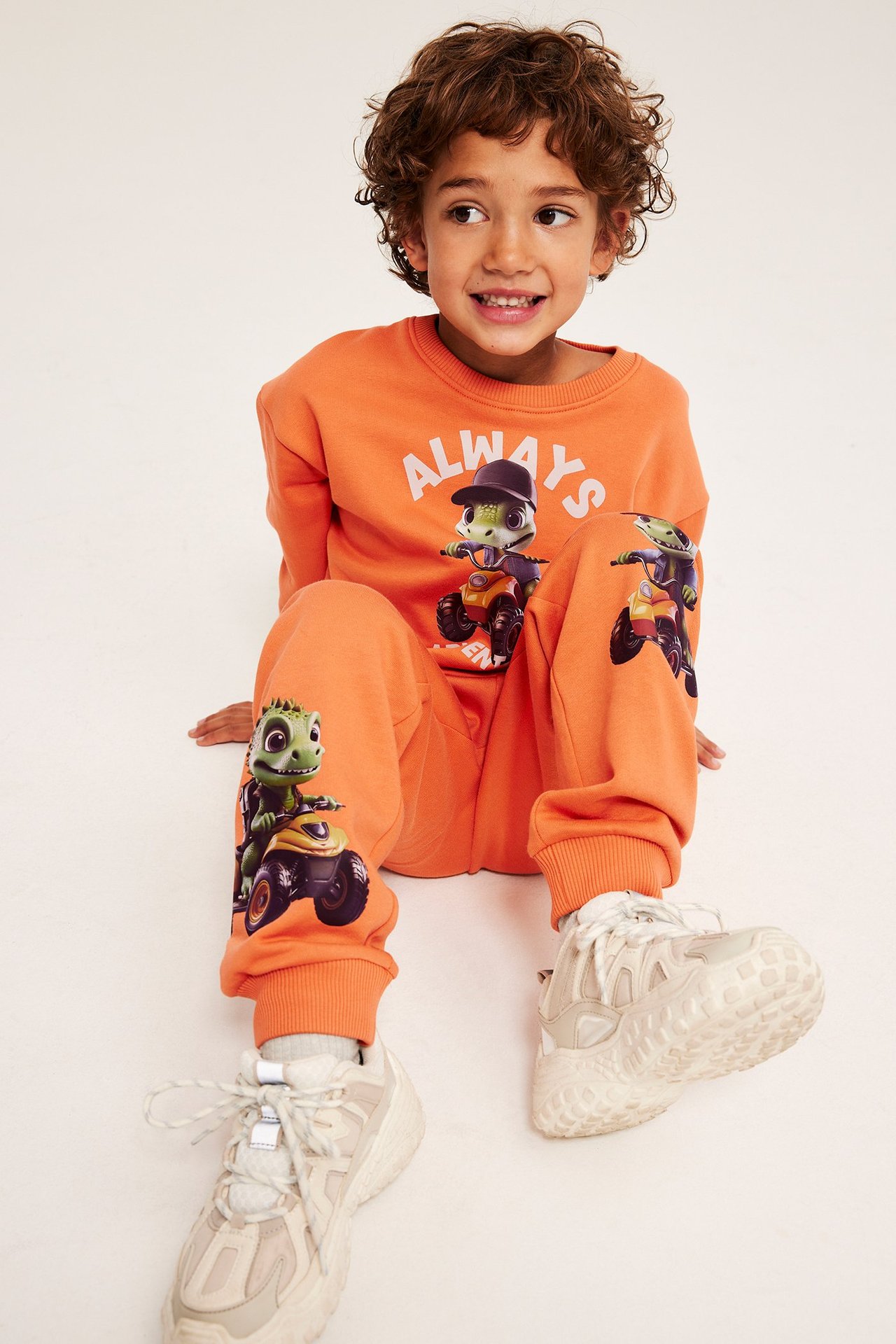 Frontalvisning: barn i oransje joggebukse med dinotrykk, matchende genser og lyse joggesko.