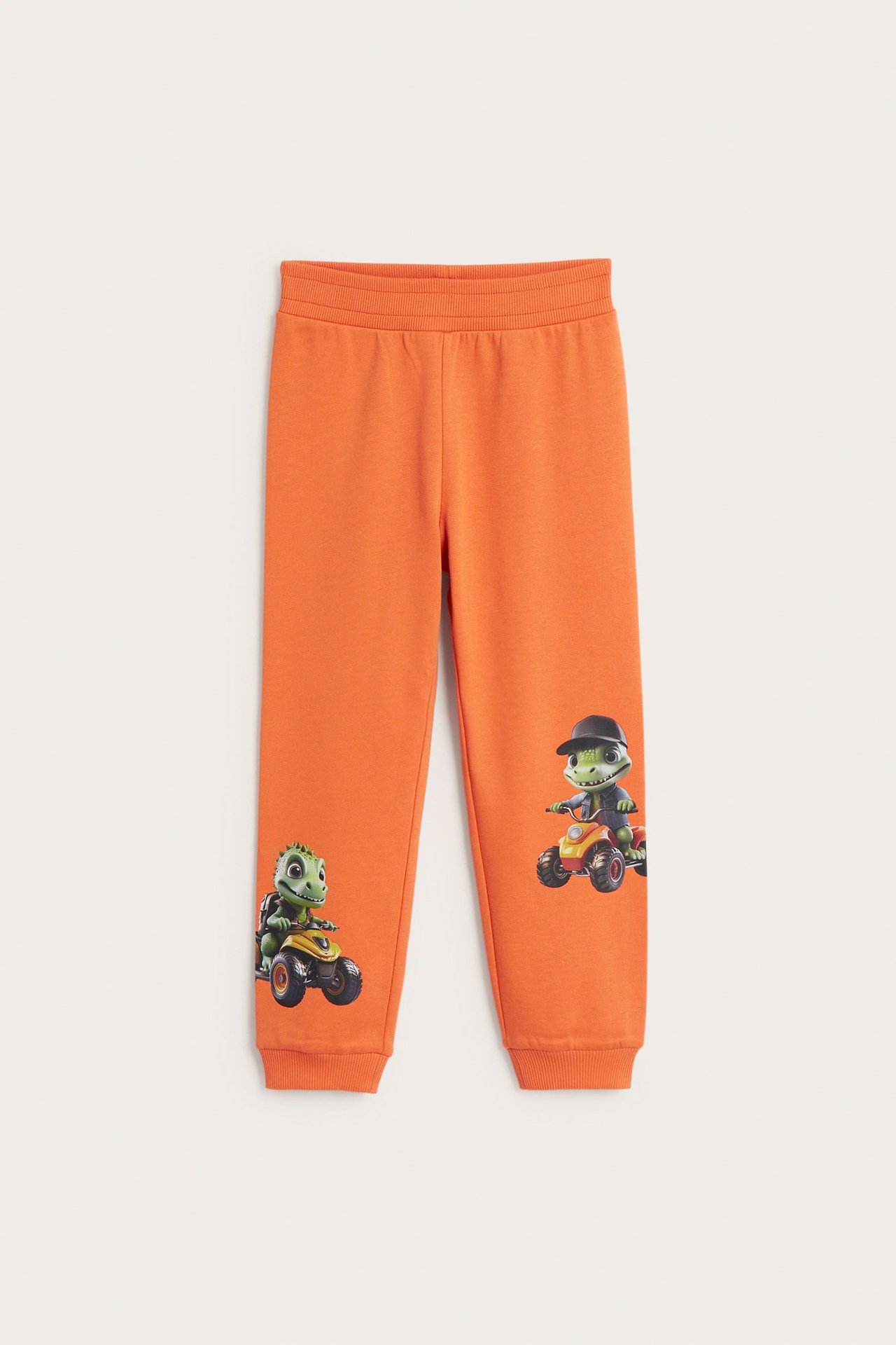 Front: oransje bomullsjoggebukse for barn med ribbet midje og dinosaurtrykk på beina.