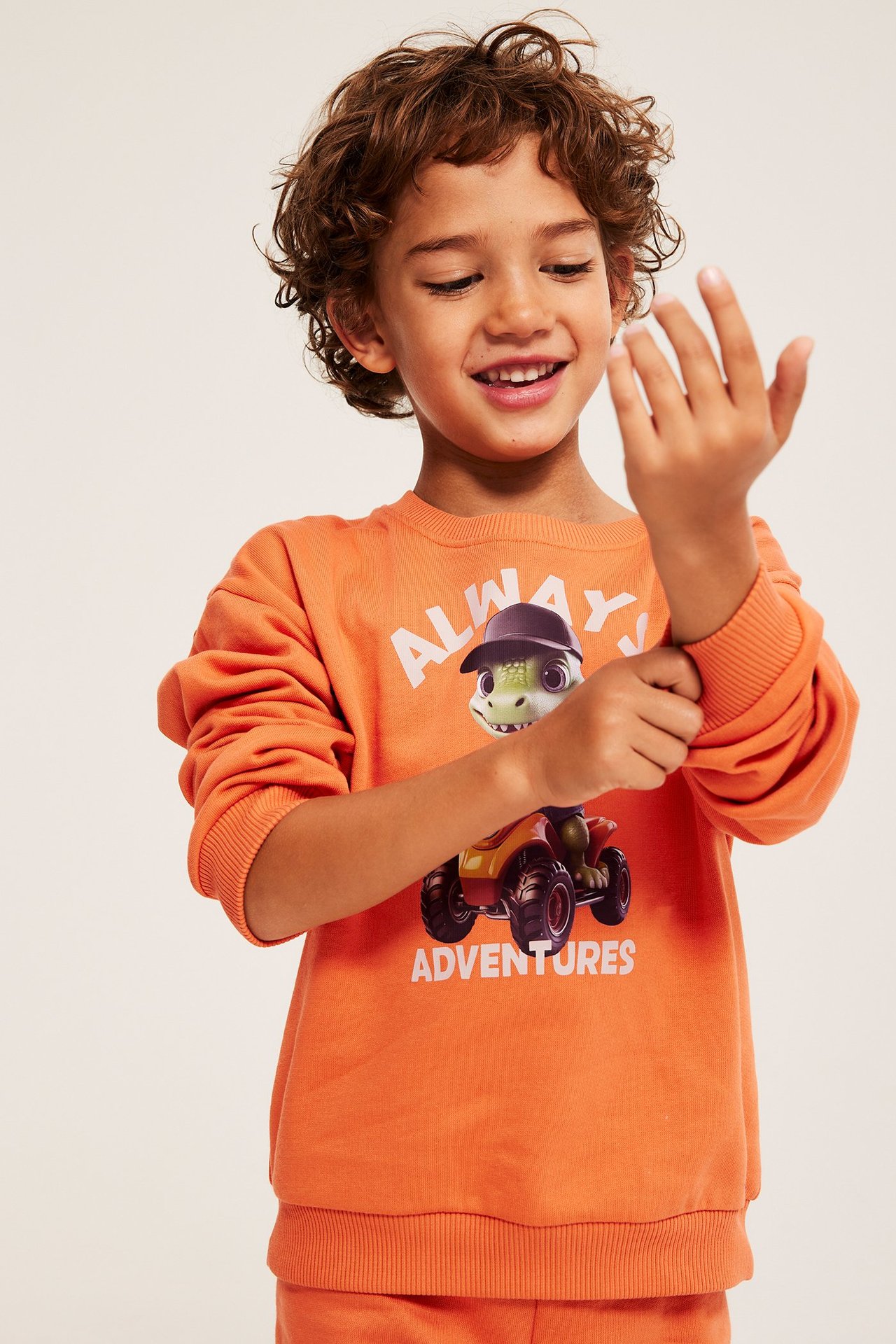 Forfra, et smilende barn i oransje bomullssweatshirt med dinosaur-firehjuling-trykk.
