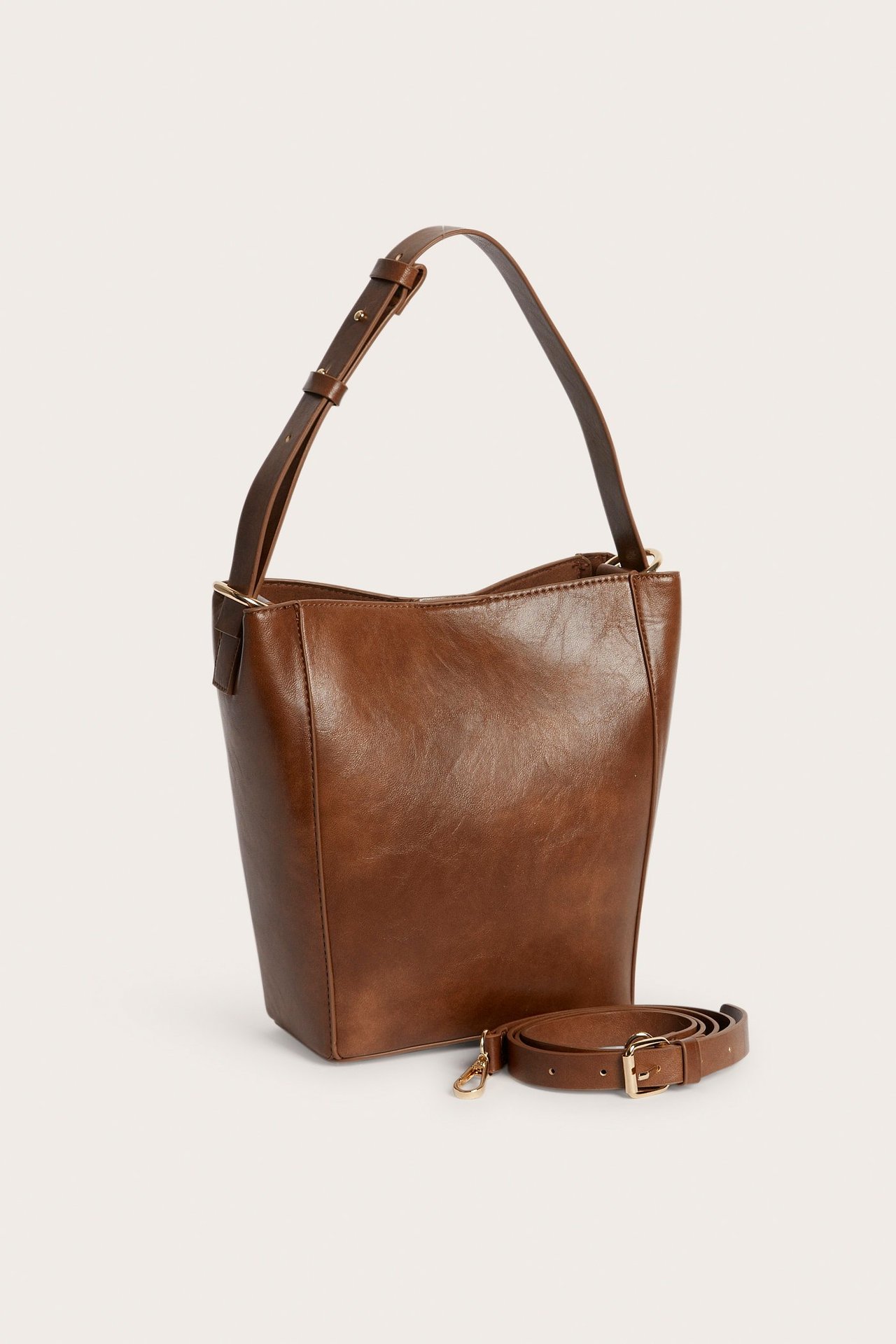 Frontvisning av en brun bøtteveske i kunstskinn med justerbar stropp, pluss en lengre stropp.