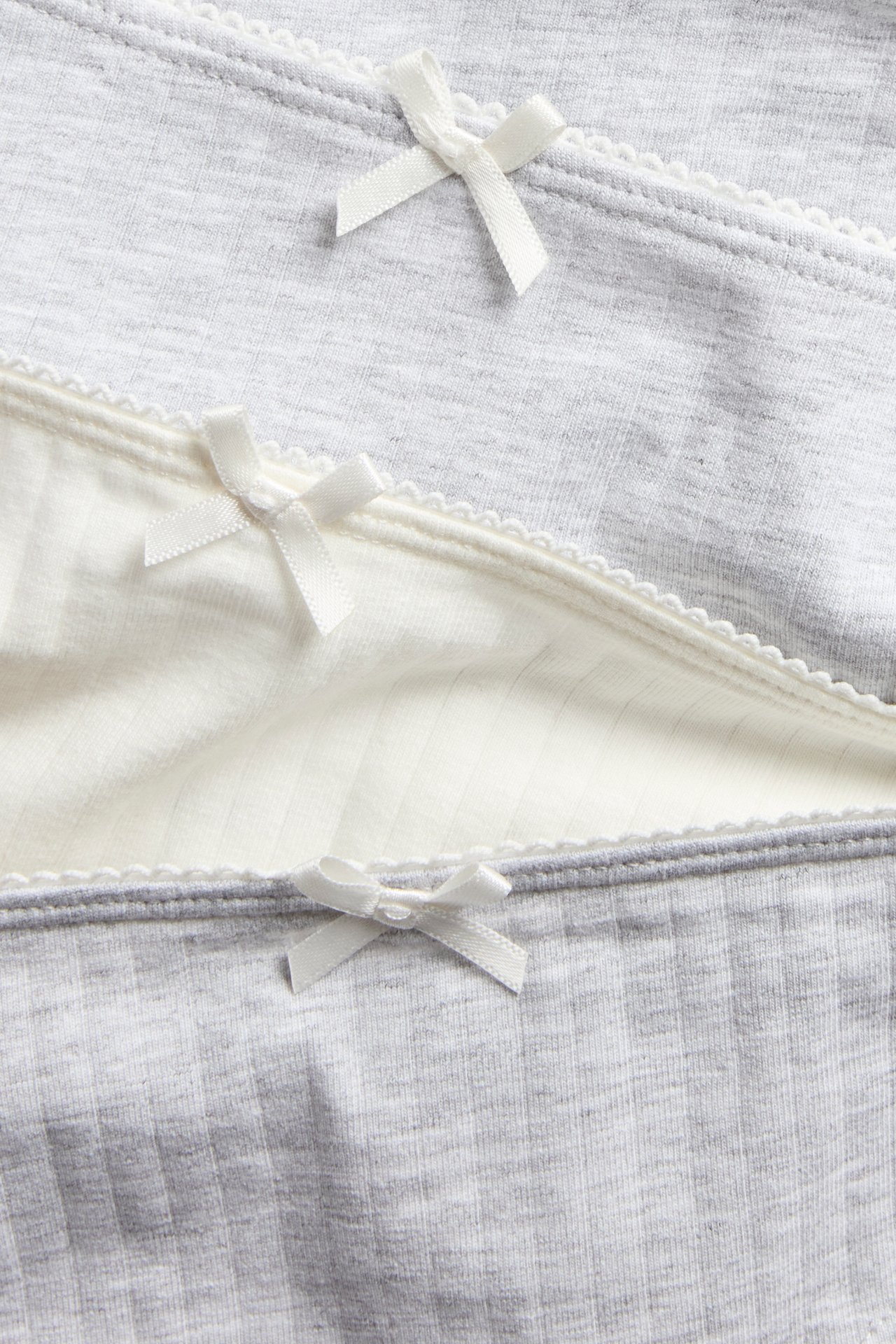 Zbliżenie: Trzy prążkowane majtki dziecięce, dwa jasnoszare, jedne ecru, z brzegami picot i kokardkami.