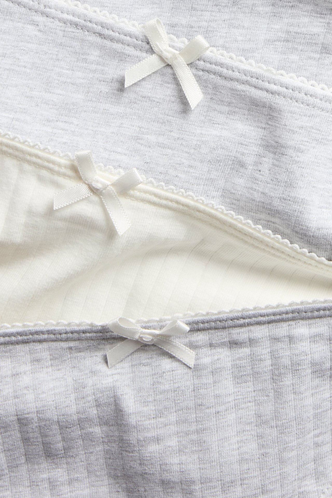 Zbliżenie: Trzy prążkowane majtki dziecięce, dwa jasnoszare, jedne ecru, z brzegami picot i kokardkami.