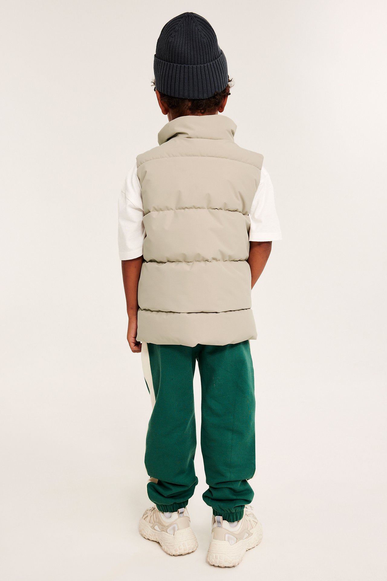 Ryggvy: Ett barn i beige pufferväst över vit t-shirt, mörkgröna mjukisbyxor, mörkgrå mössa och beiga sneakers.