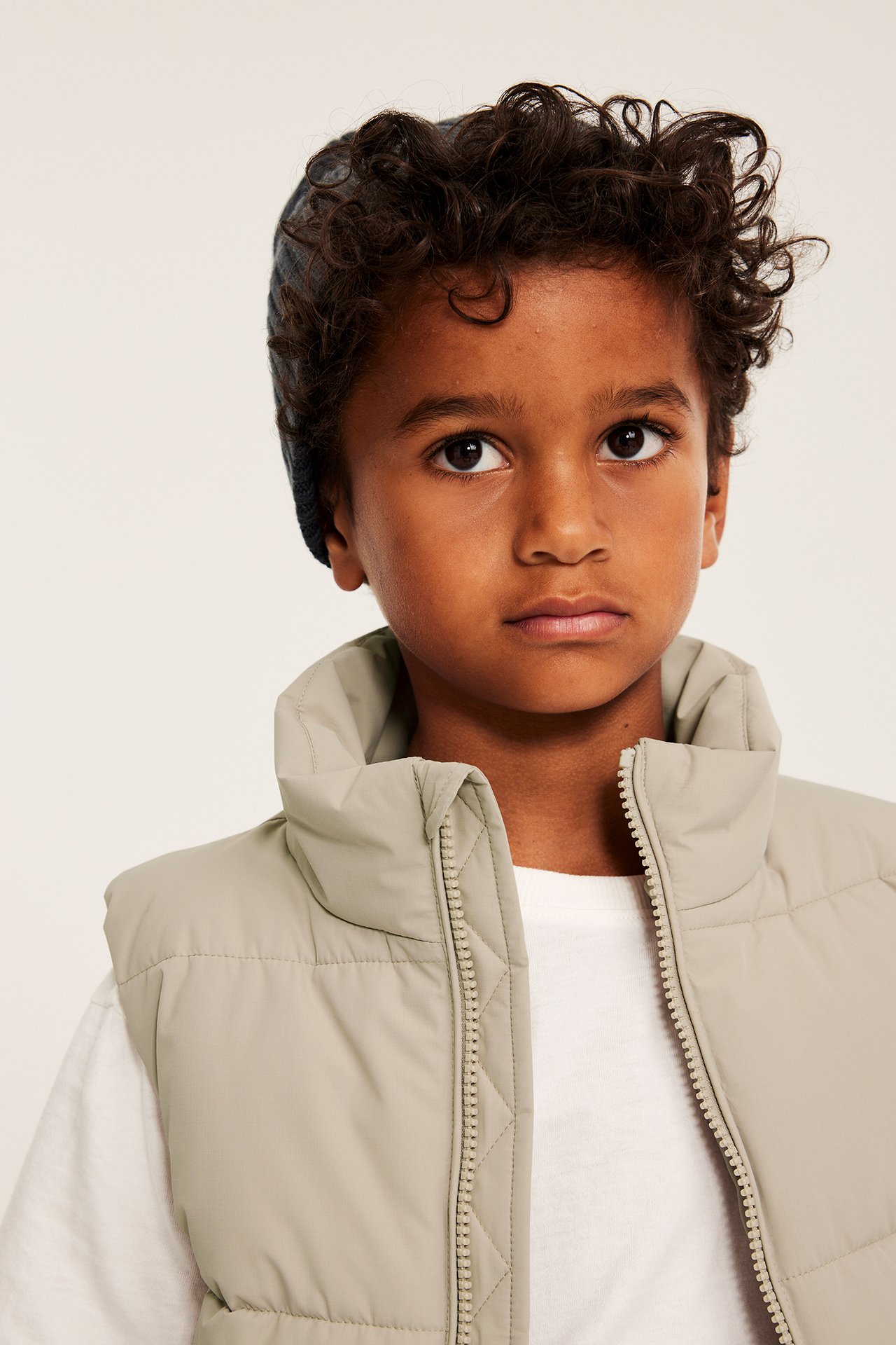 Edestäpäin: Lapsi beigessä pehmustetussa liivissä, korkea kaulus, vetoketju, ja tumma pipo.