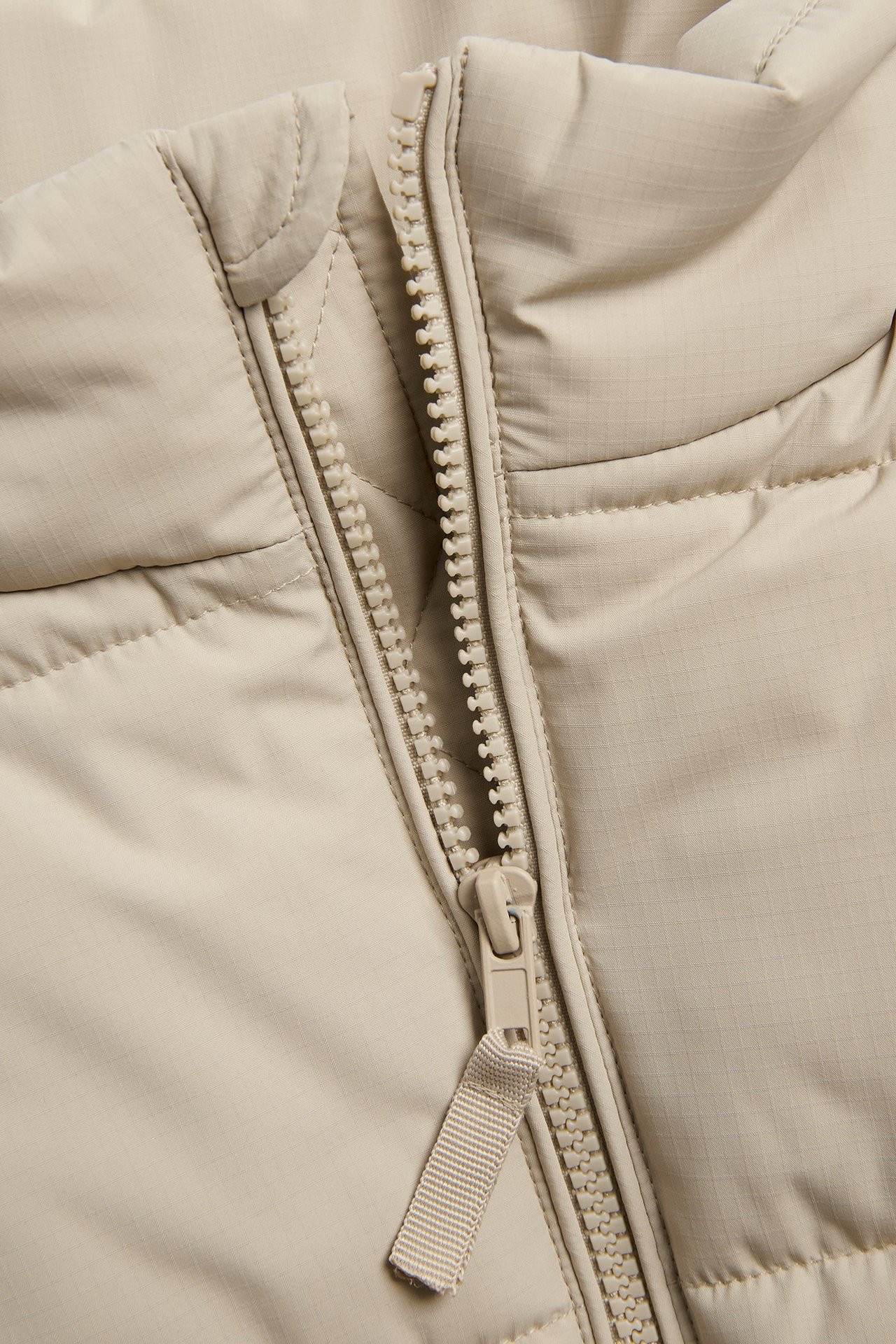 Zbliżenie na beżową, pikowaną kamizelkę puchową z częściowo rozpiętym zamkiem z przodu i przeszyciami.